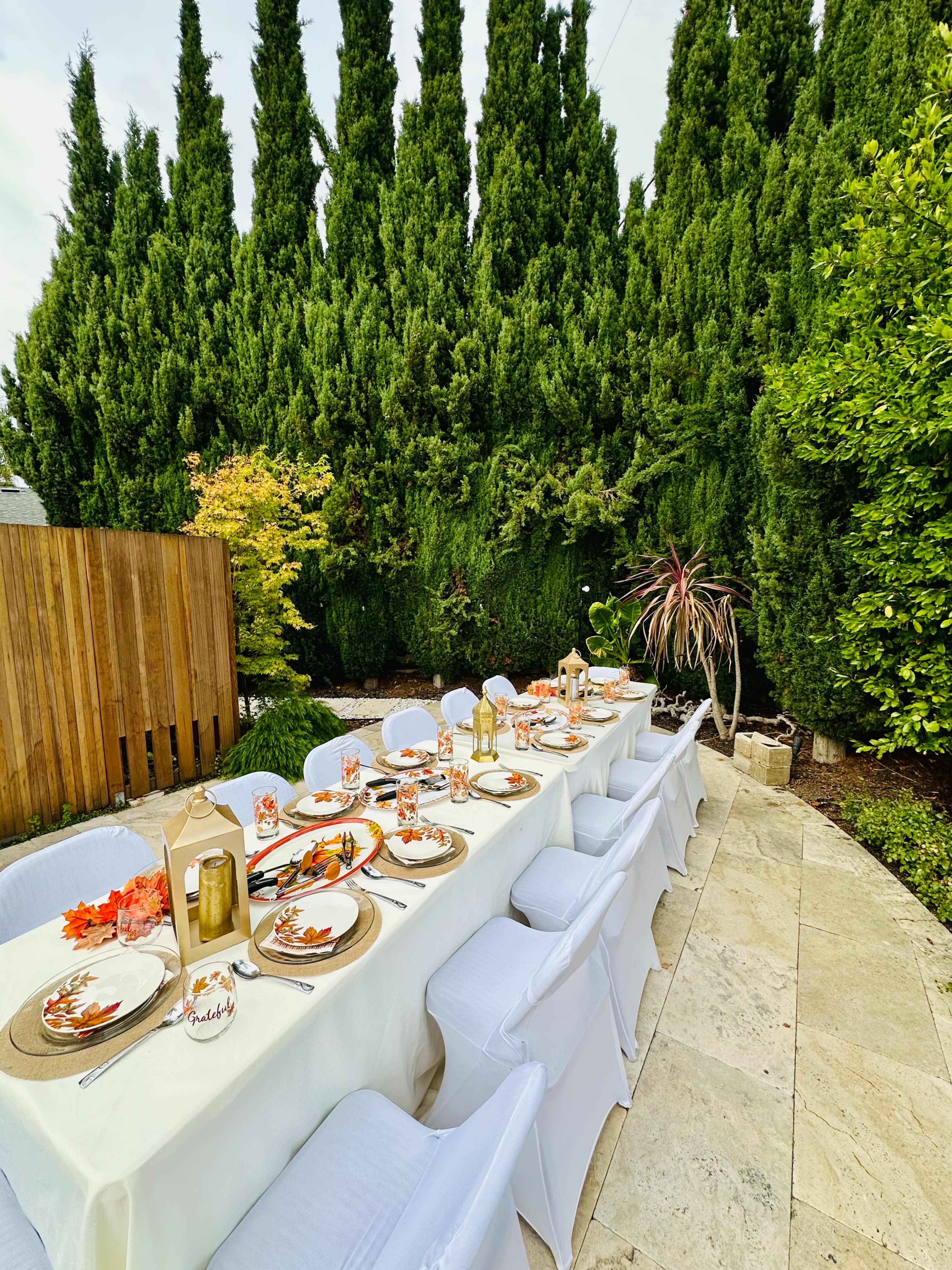 A long dining table is set outdoors with white tablecloth and chairs, surrounded by lush greenery and featuring decorative centerpieces.