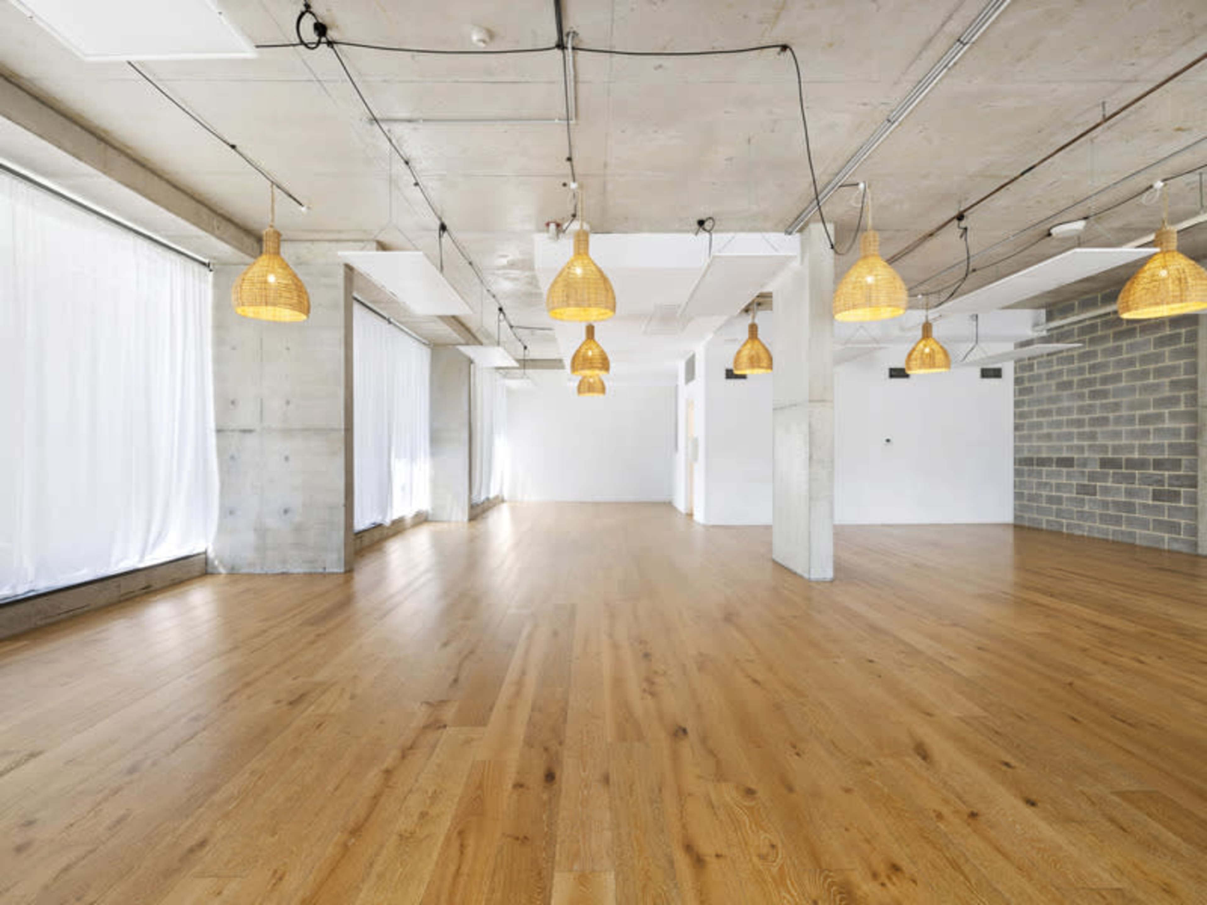 The image shows a spacious, empty room with wooden floors, large windows dressed in white curtains, and pendant lights hanging from the ceiling.