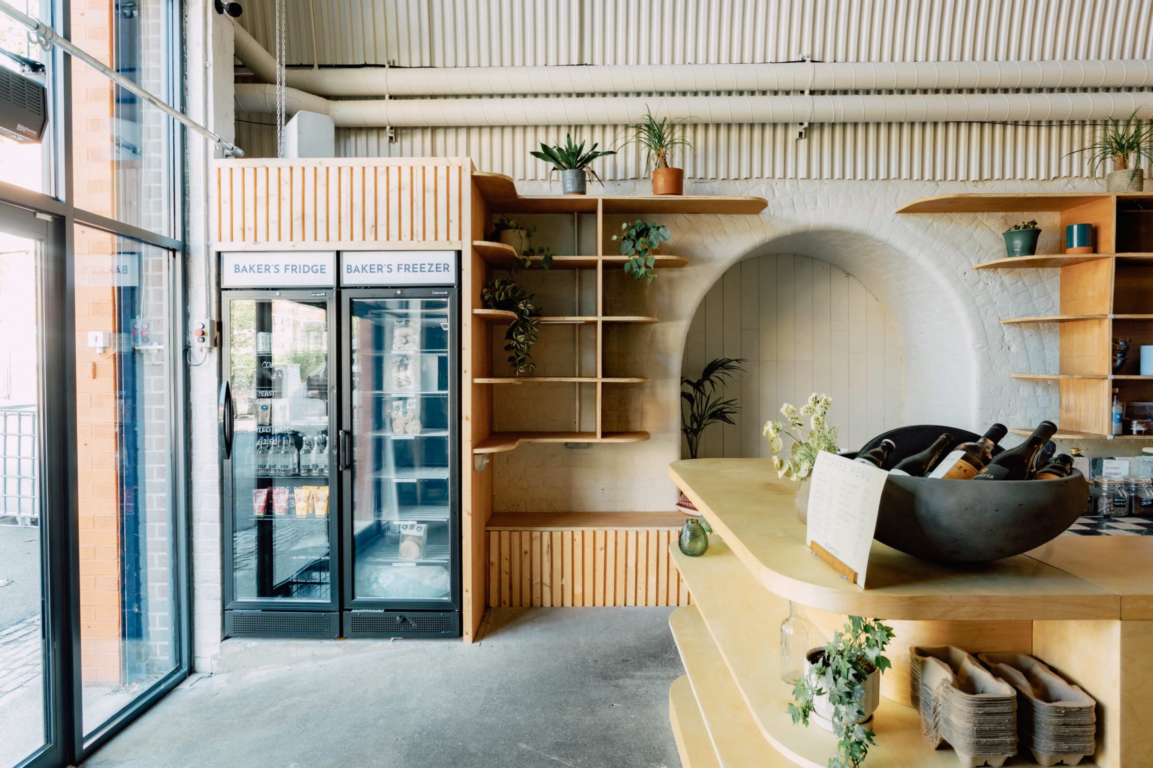 The image shows a bright, modern bakery interior featuring a wooden counter, shelves with plants, and a glass fridge displaying baked goods.