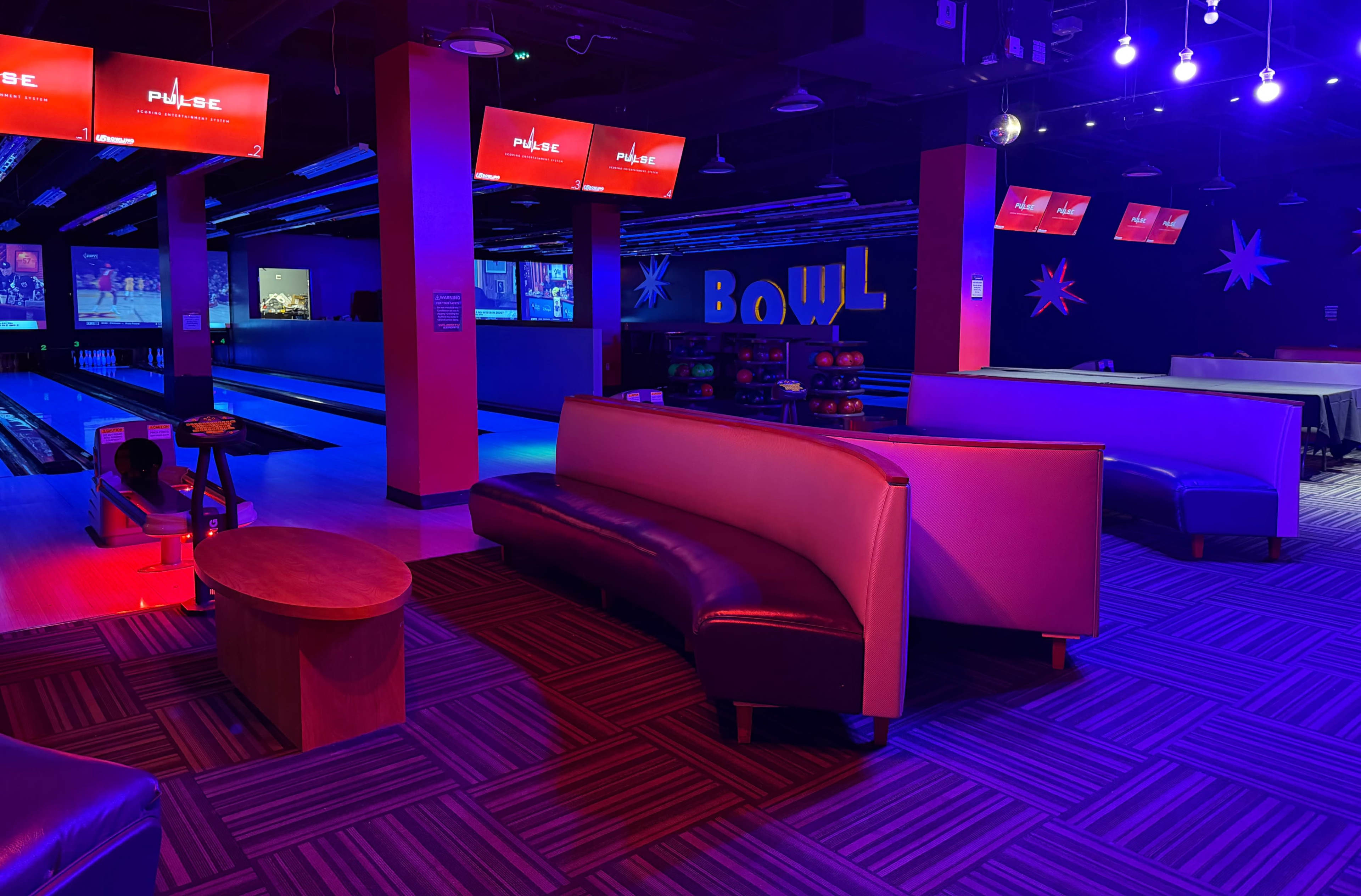 The image shows a bowling alley interior with seating areas, illuminated by colorful lights and multiple screens displaying the word "PULSE."