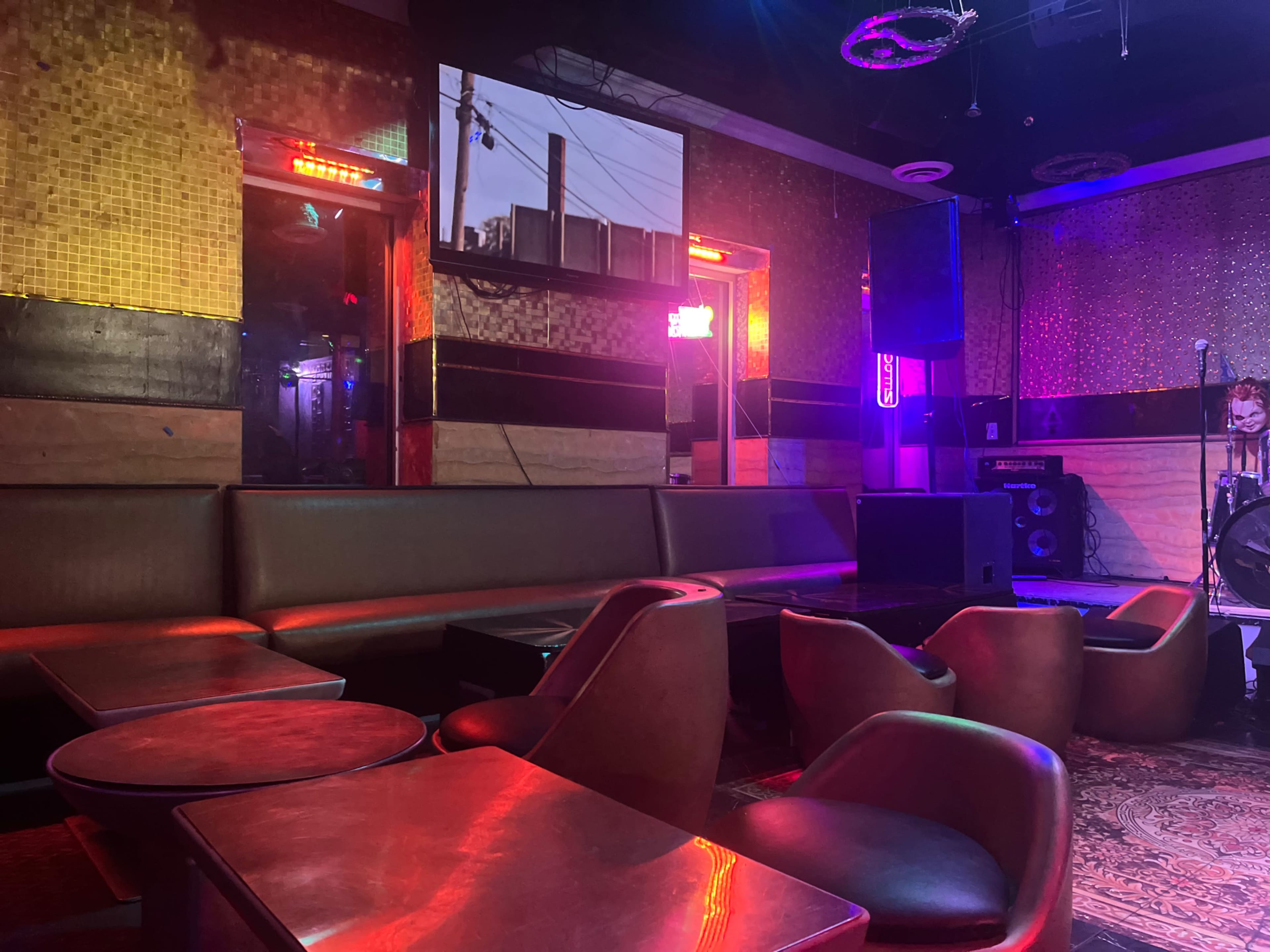 The image shows a dimly lit lounge area with brown leather seating, small round tables, and colorful neon lights illuminating the space.