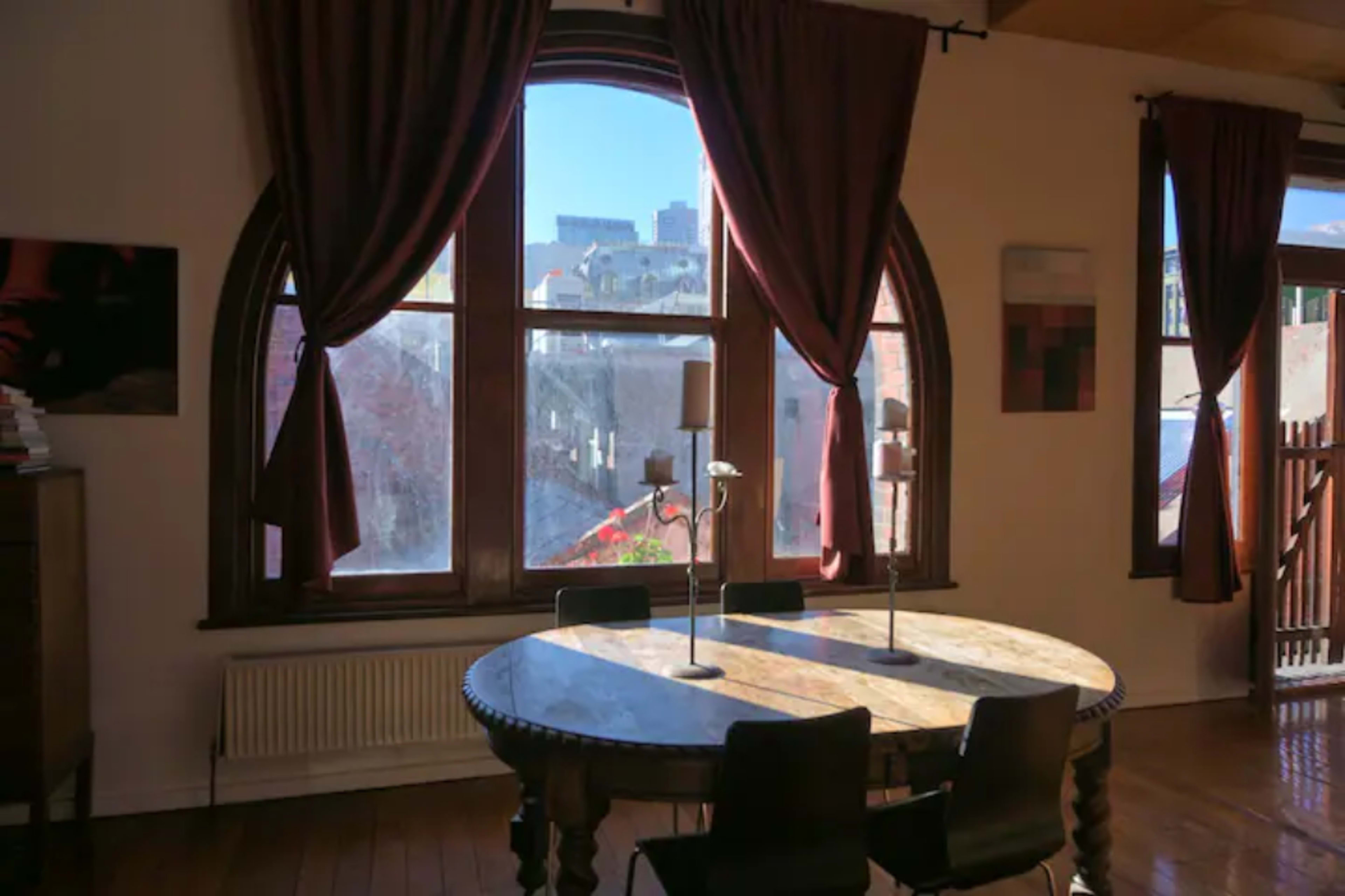 A round wooden table with two lamps stands in a room featuring large arched windows dressed with dark curtains, revealing a view of an urban landscape outside.