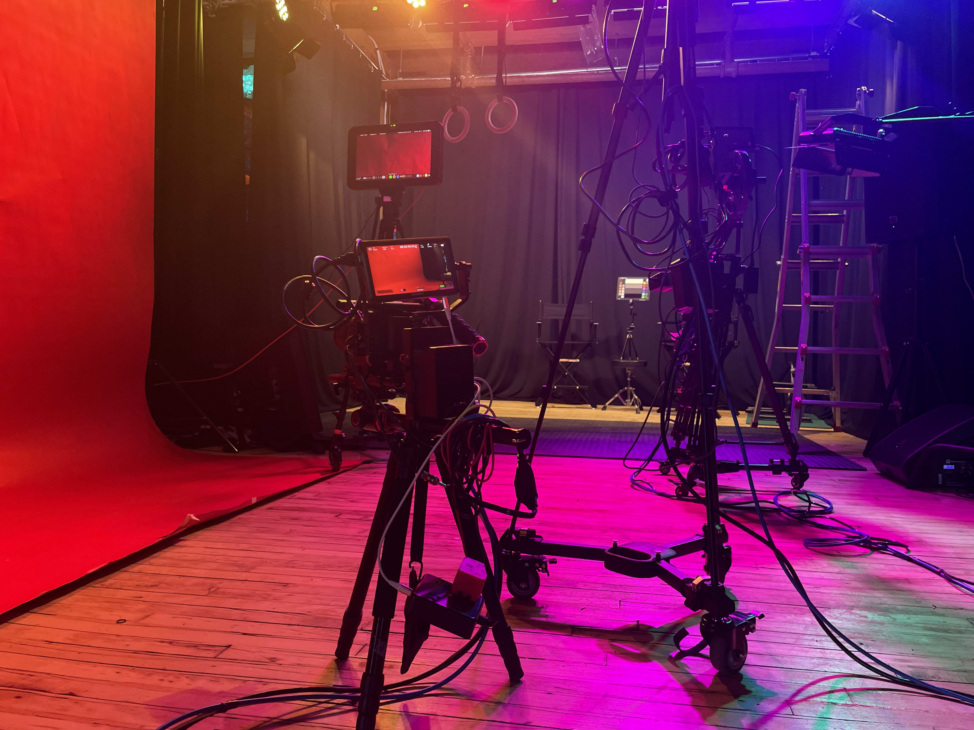 The scene shows a partially set up studio space with vibrant lighting, tripods, and audio-visual equipment on a wooden floor.