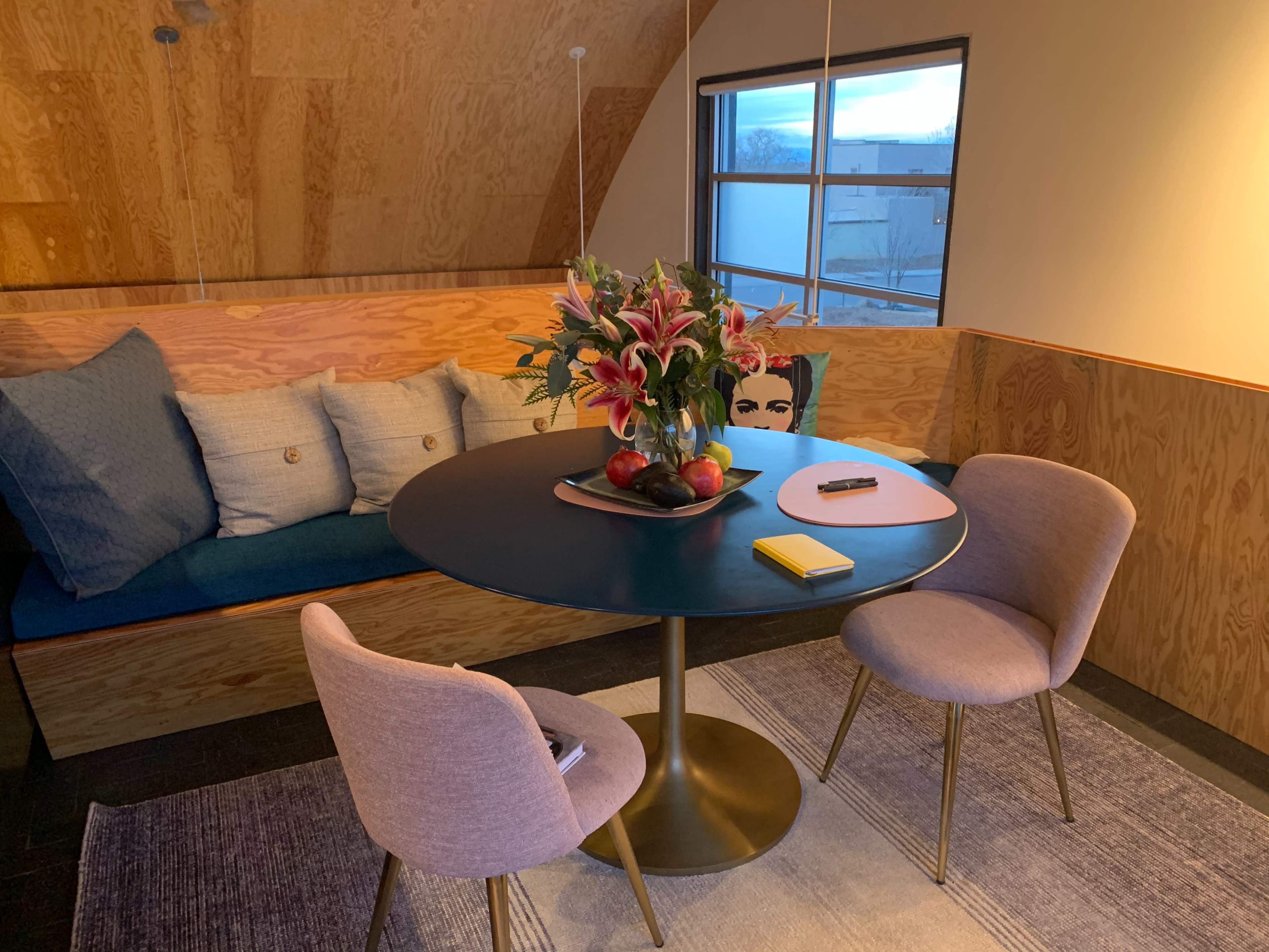 A round table with two pink chairs is set near a sofa adorned with blue cushions and a bouquet of flowers, alongside a bowl of apples and a notepad.