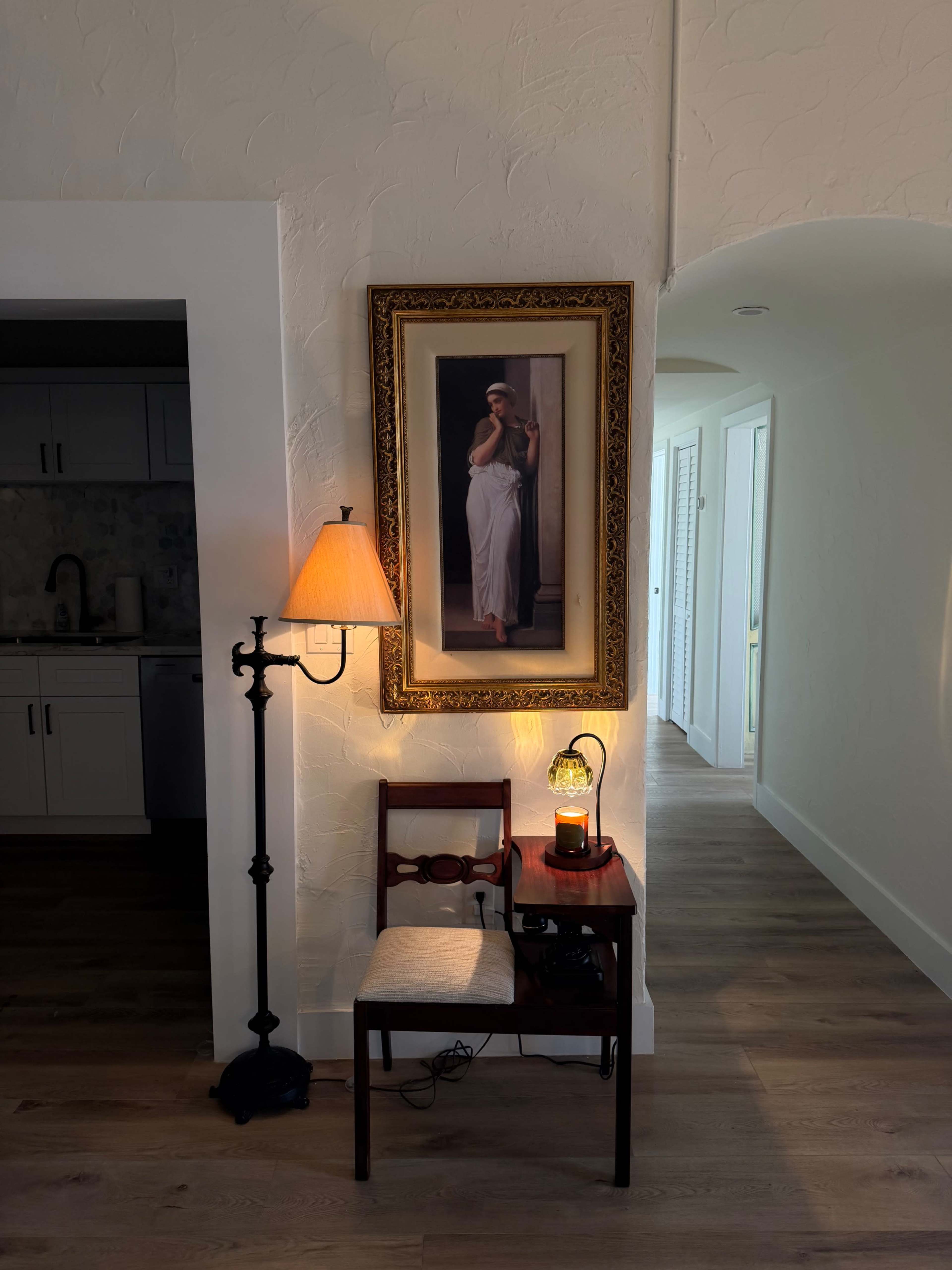 A wooden chair and small table are situated beside a lamp and a framed painting in a brightly lit hallway.