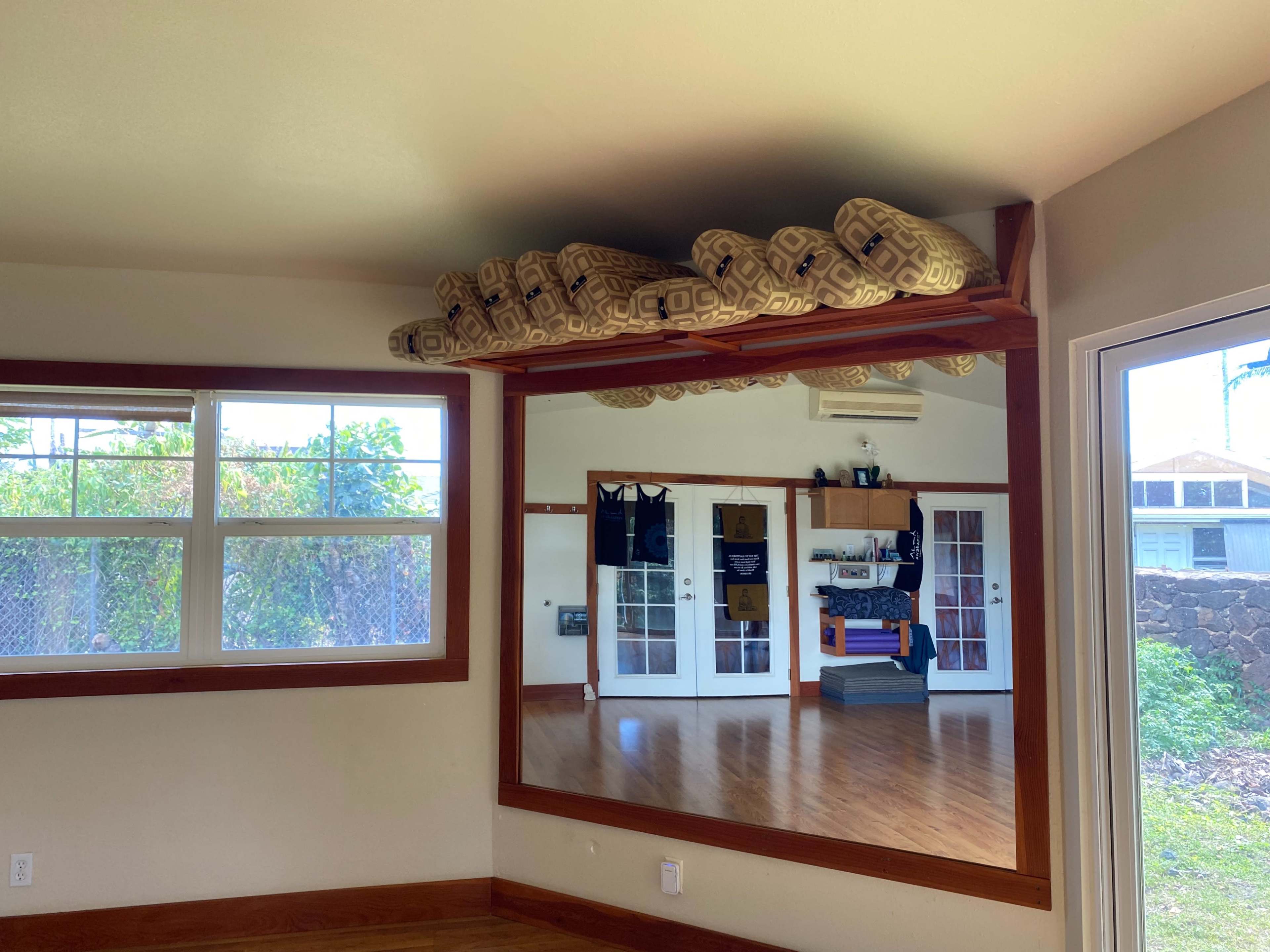 A room with hardwood floors, a large mirror on one wall, and a shelf above with patterned cushions.