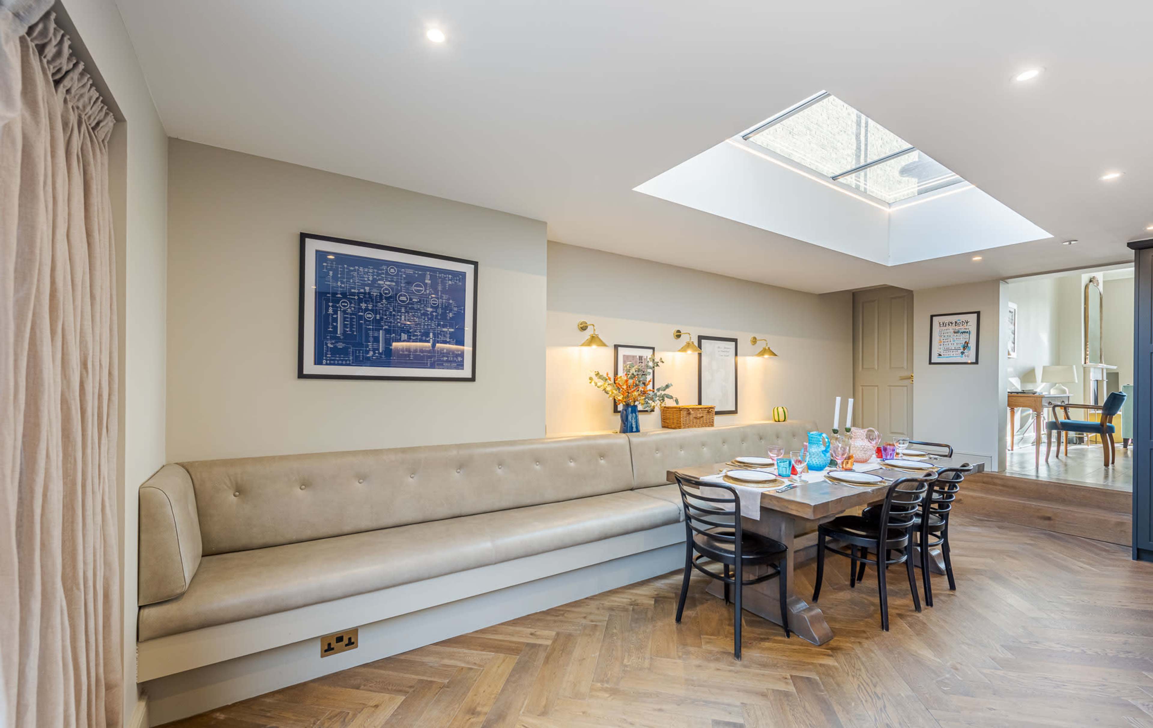 The image shows a modern dining area featuring a long upholstered bench, a wooden dining table with seating, and a skylight above illuminating the space.