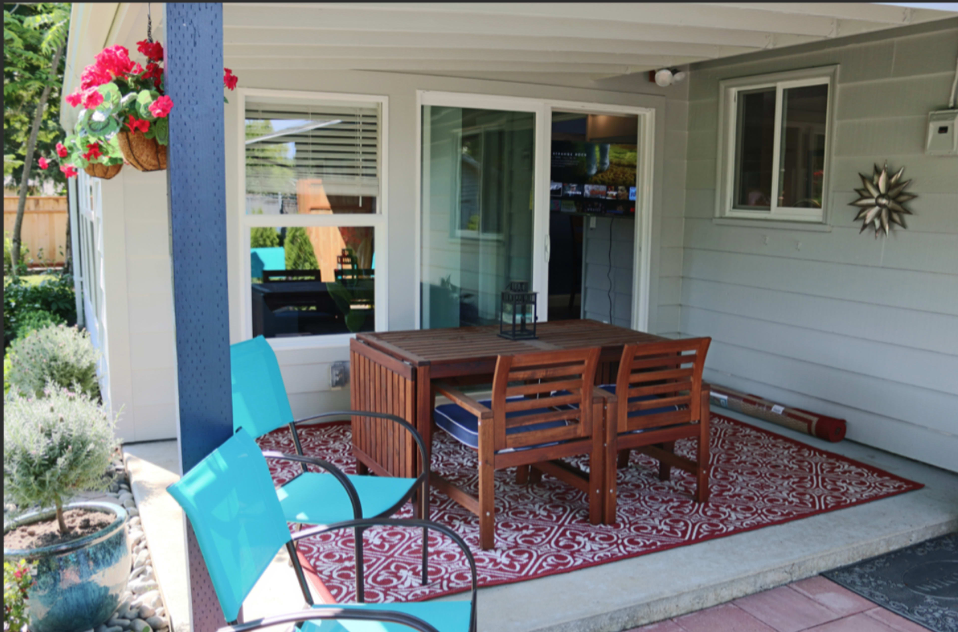 A covered patio area featuring a wooden dining table with four chairs on a patterned rug, surrounded by potted plants and a television visible indoors.