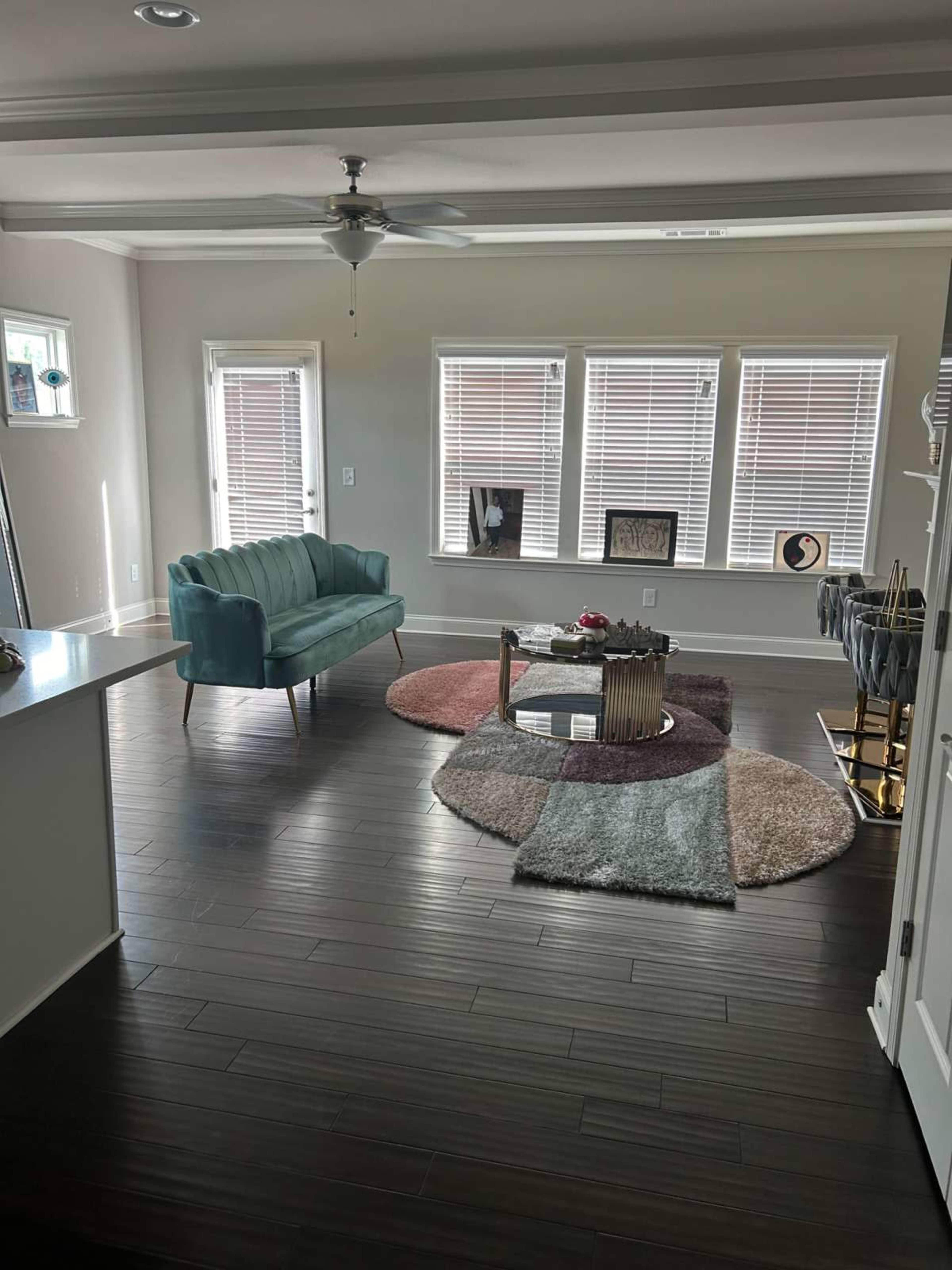 The image shows a modern living room with a teal sofa, a round coffee table, and several windows framed by white blinds, all set on dark wood flooring.
