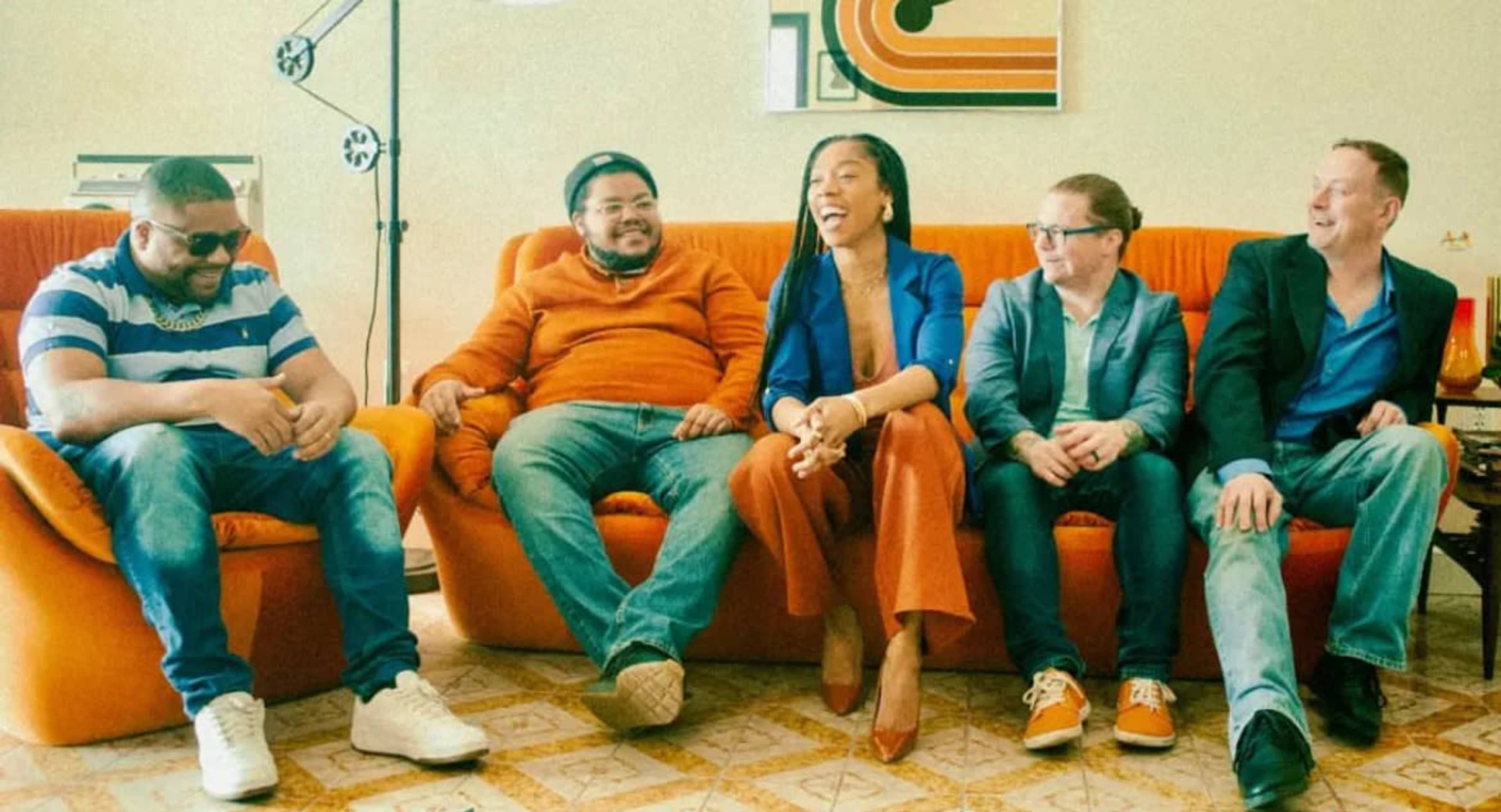 A group of five people sits on an orange couch, engaged in conversation, with various expressions on their faces.