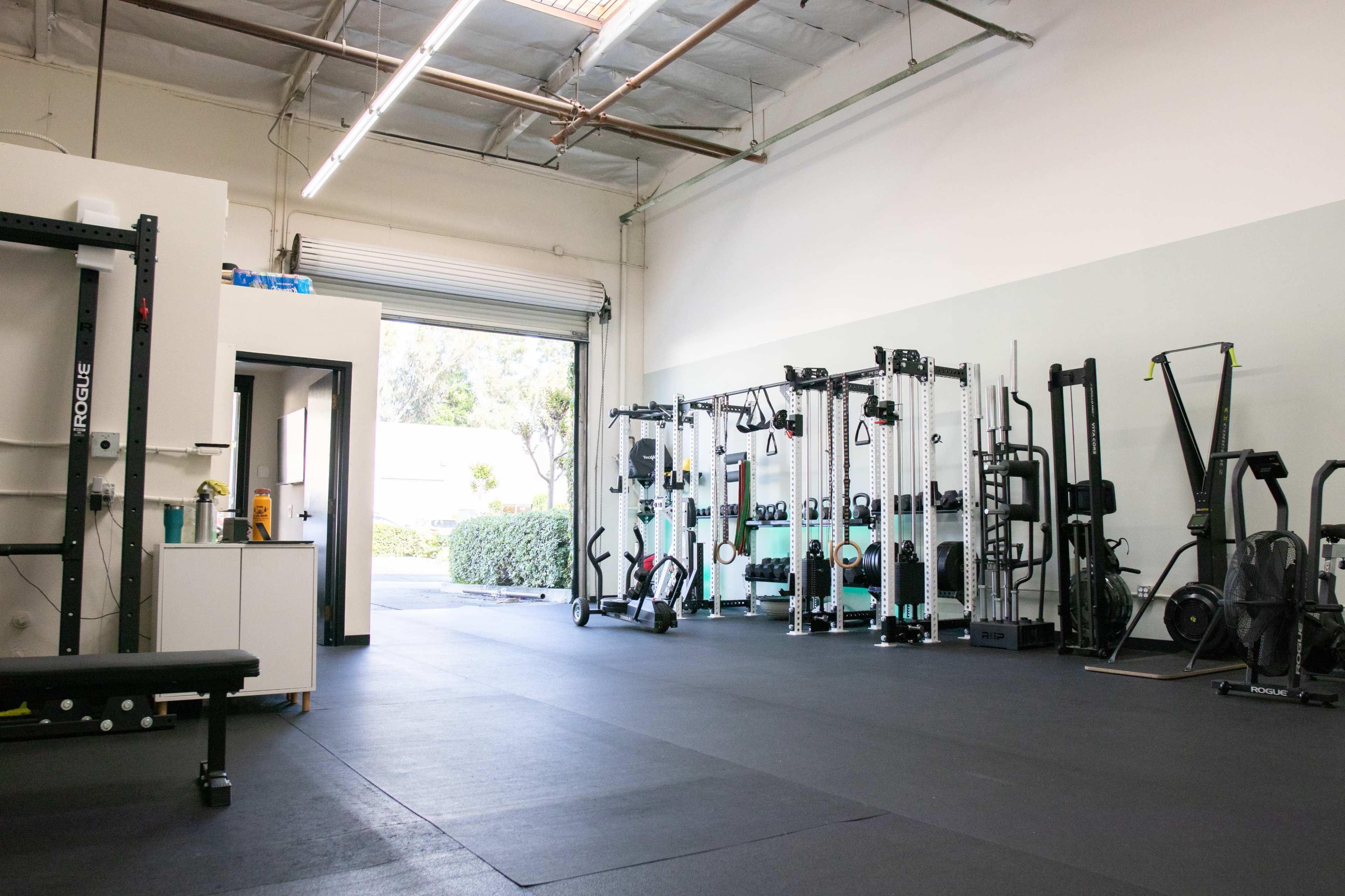 A spacious gym interior with various exercise equipment arranged along the walls and an open garage door leading to an outdoor area.