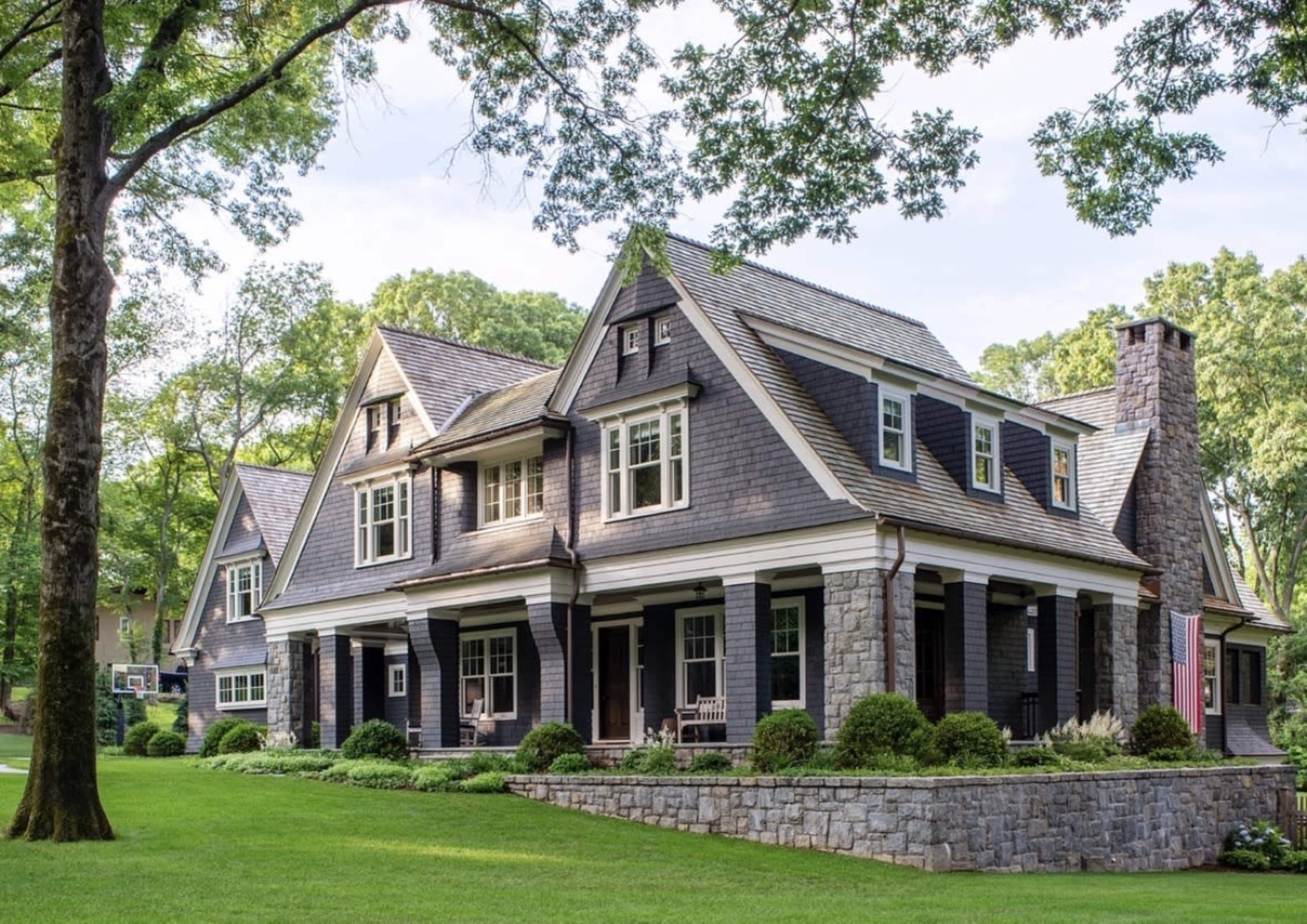The image shows a large, two-story house with a mix of stone and dark wooden siding, set in a lush green landscape with trees and a well-maintained lawn.