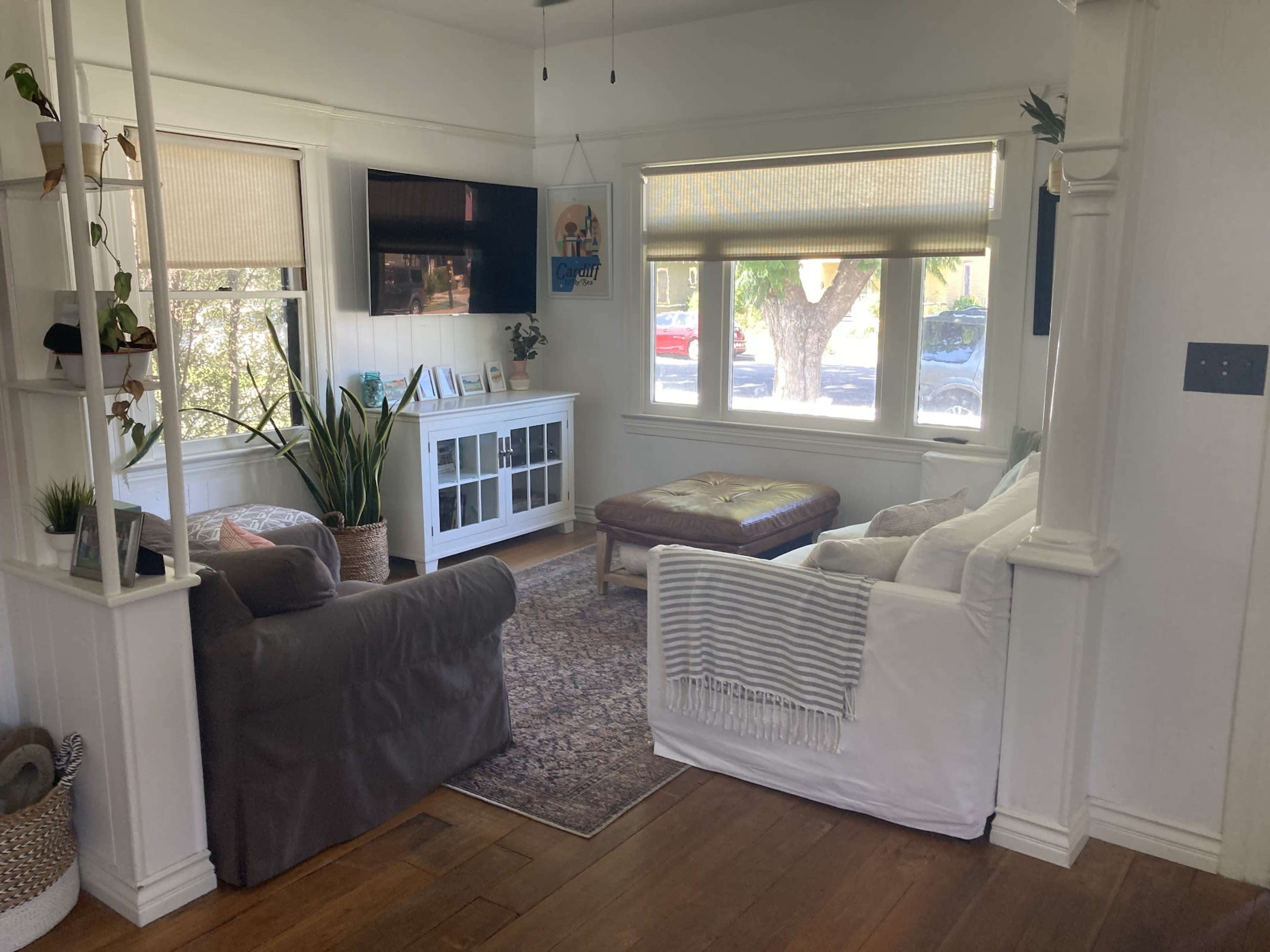 A living room with a grey armchair, a white couch, a coffee table, and a television mounted on the wall, all situated near large windows with blinds.