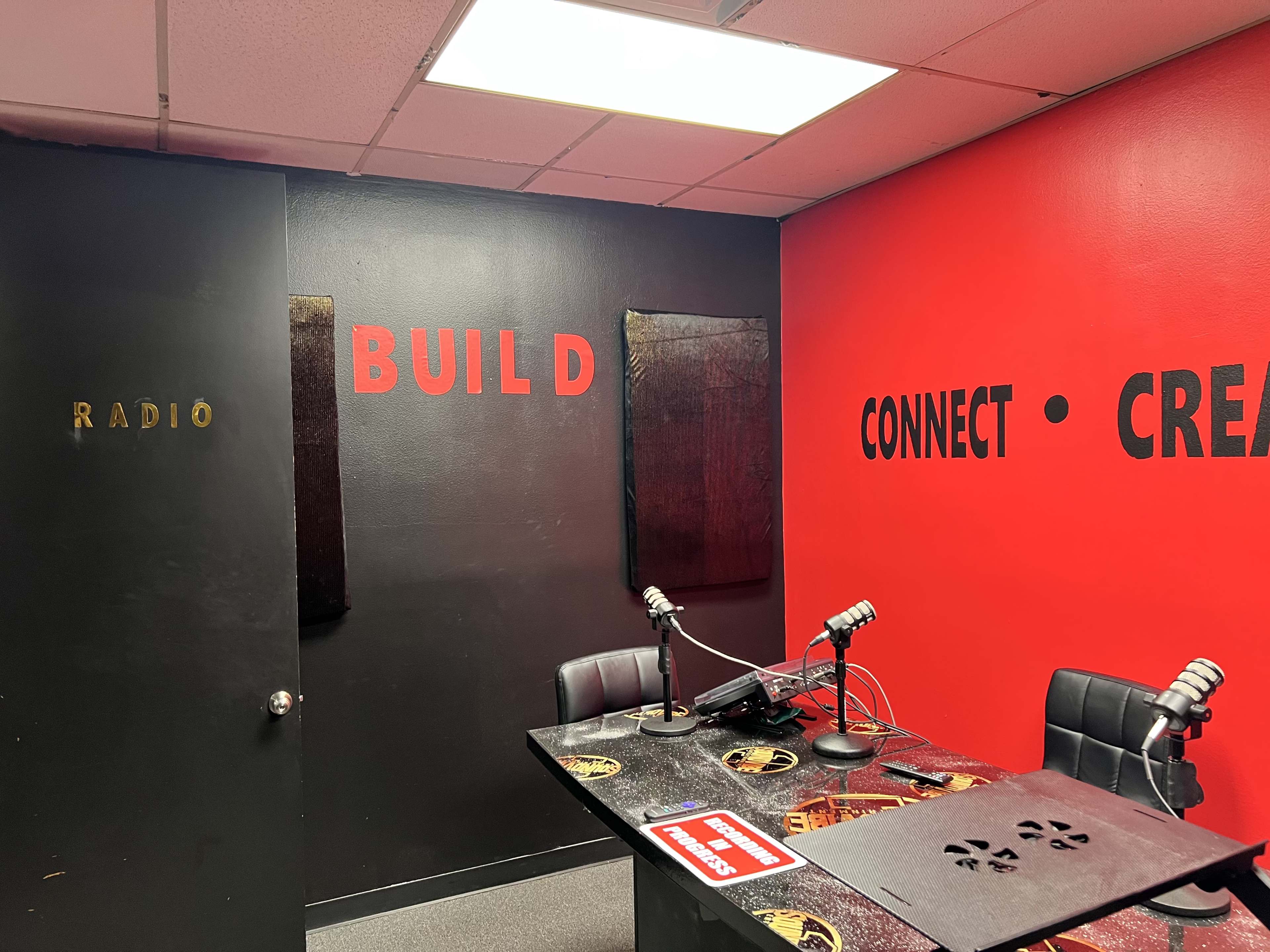 A small radio studio with red and black walls, a desk with microphones, and the words "BUILD," "CONNECT," and "CREATE" displayed on the wall.