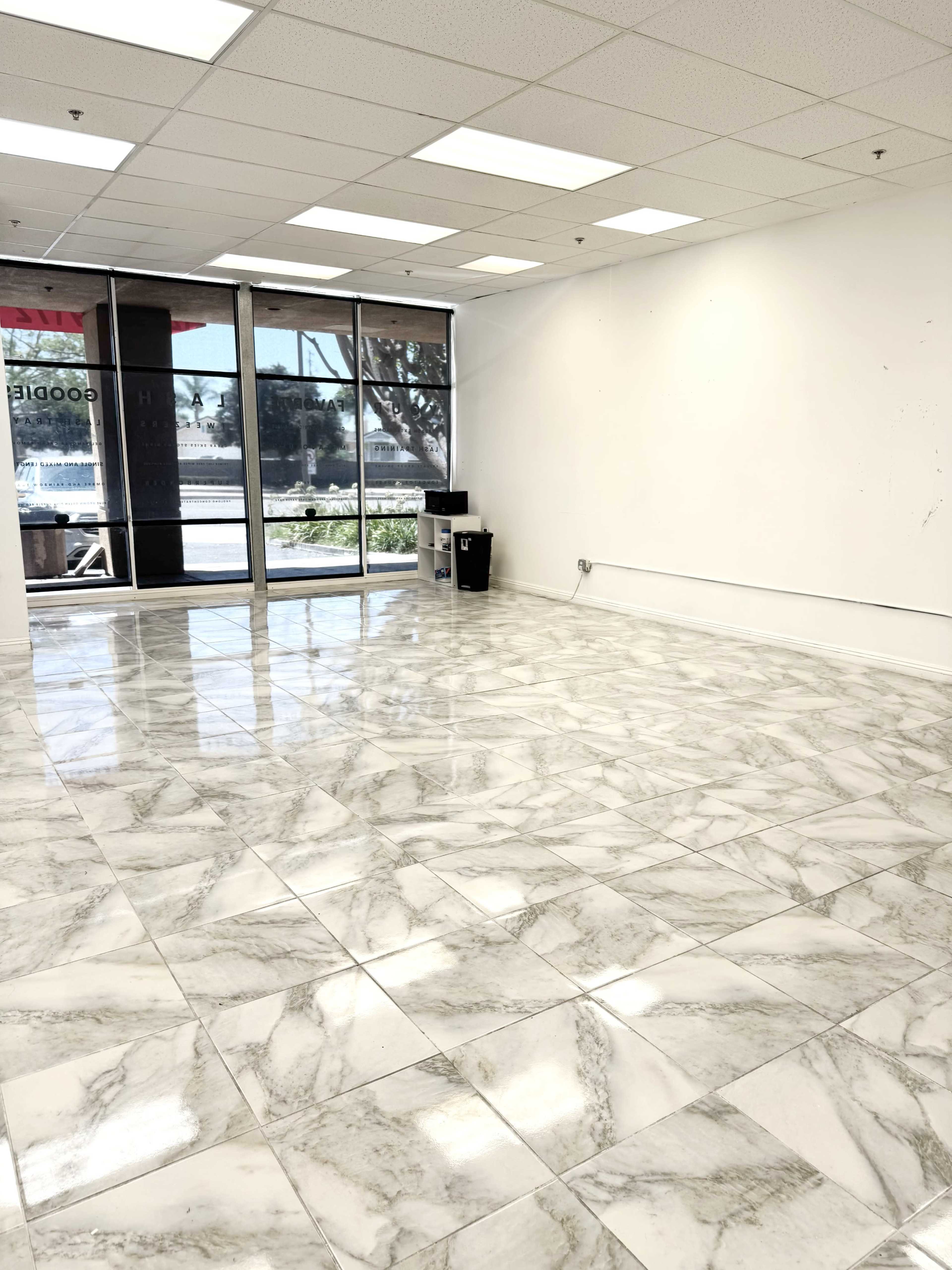The image shows a spacious, empty room with marble-patterned tile flooring and large windows along one wall allowing natural light to enter.
