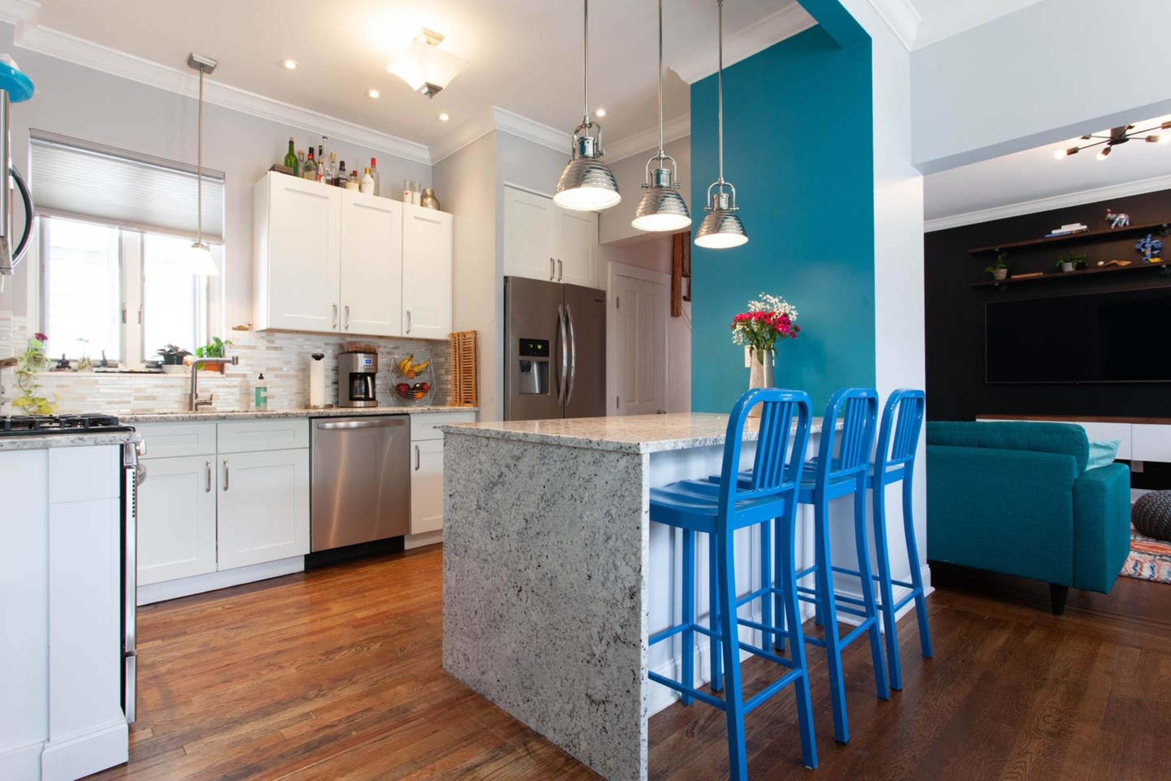 A modern kitchen features white cabinets, stainless steel appliances, and a blue accent wall with three blue bar stools at a stone island.