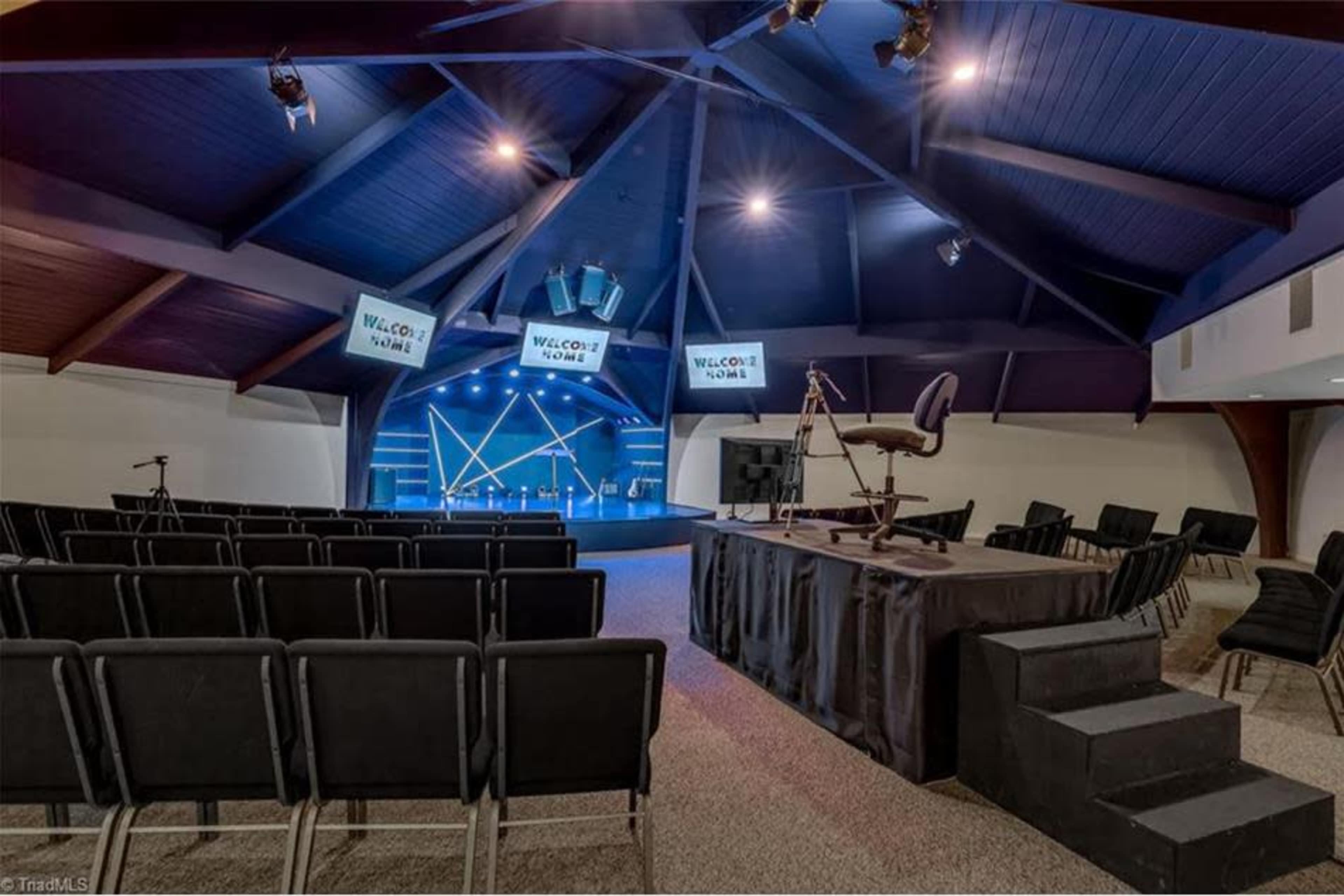 A spacious auditorium with rows of black chairs, a stage equipped with lighting, and several screens displaying a welcome message.