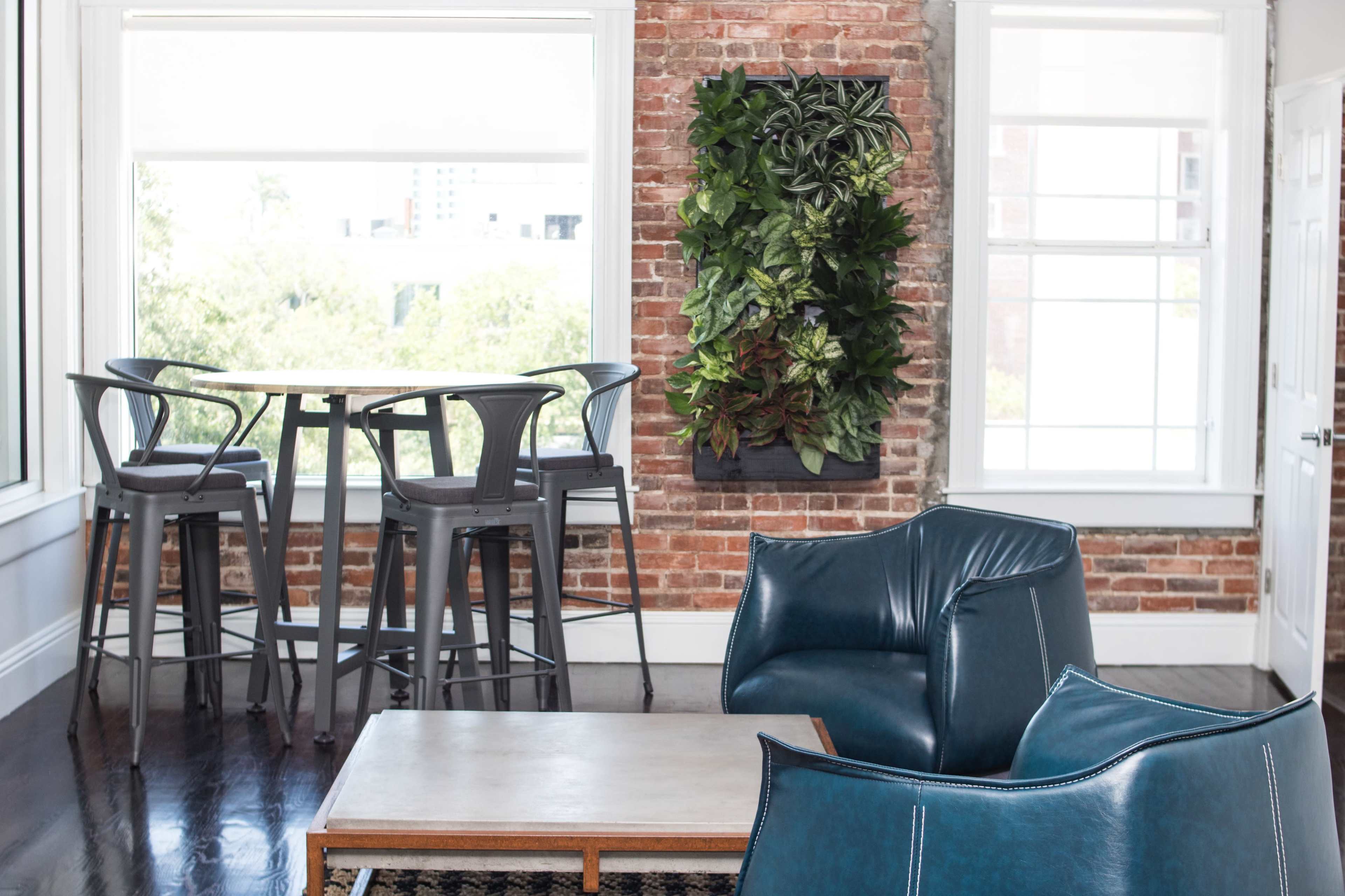 The image shows a modern interior space featuring a brick wall, a small dining table with high chairs, a vertical plant installation, and a blue armchair.