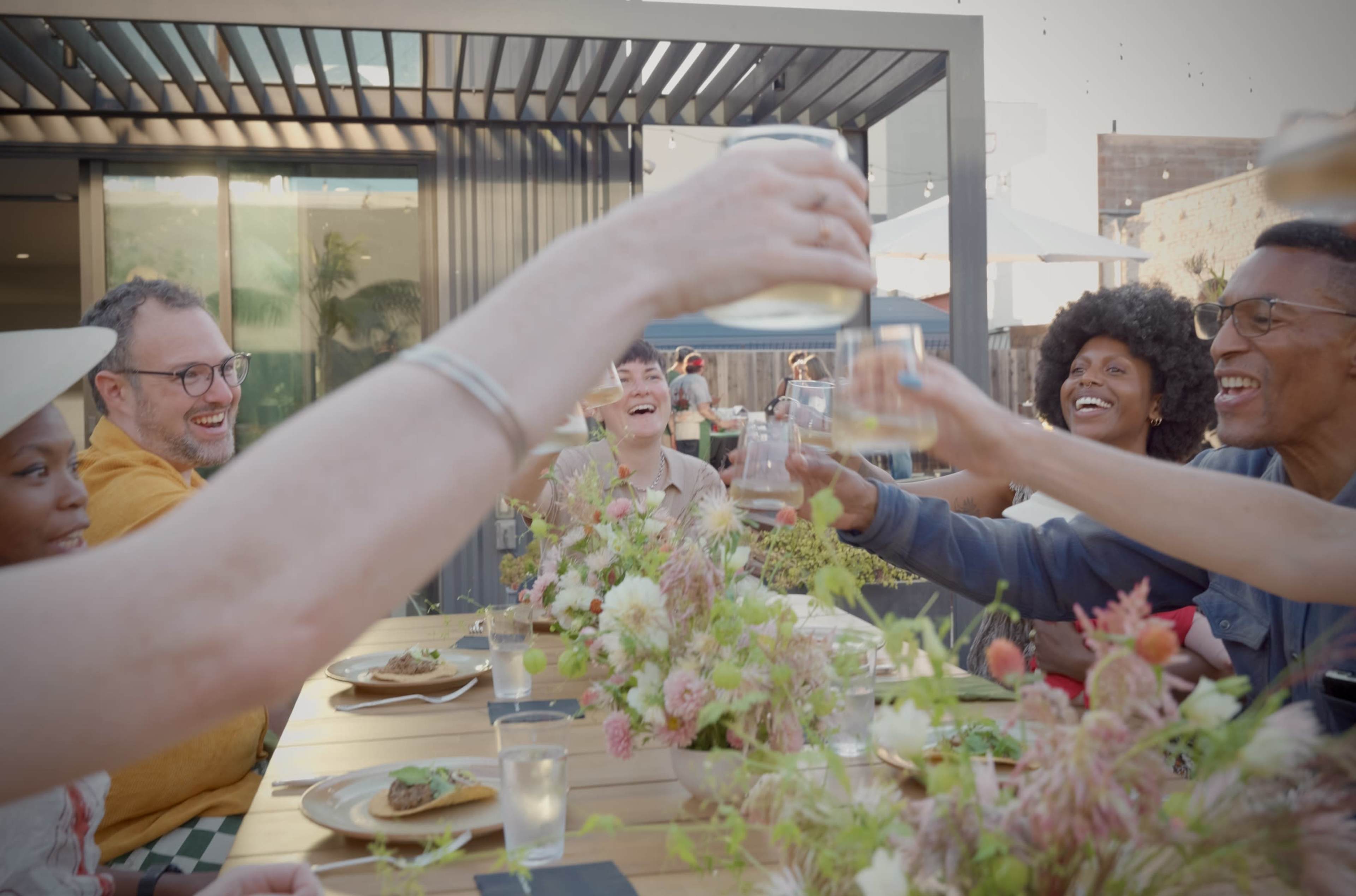 A group of people are gathered around a table, raising their glasses in a toast while enjoying a meal among floral decorations.