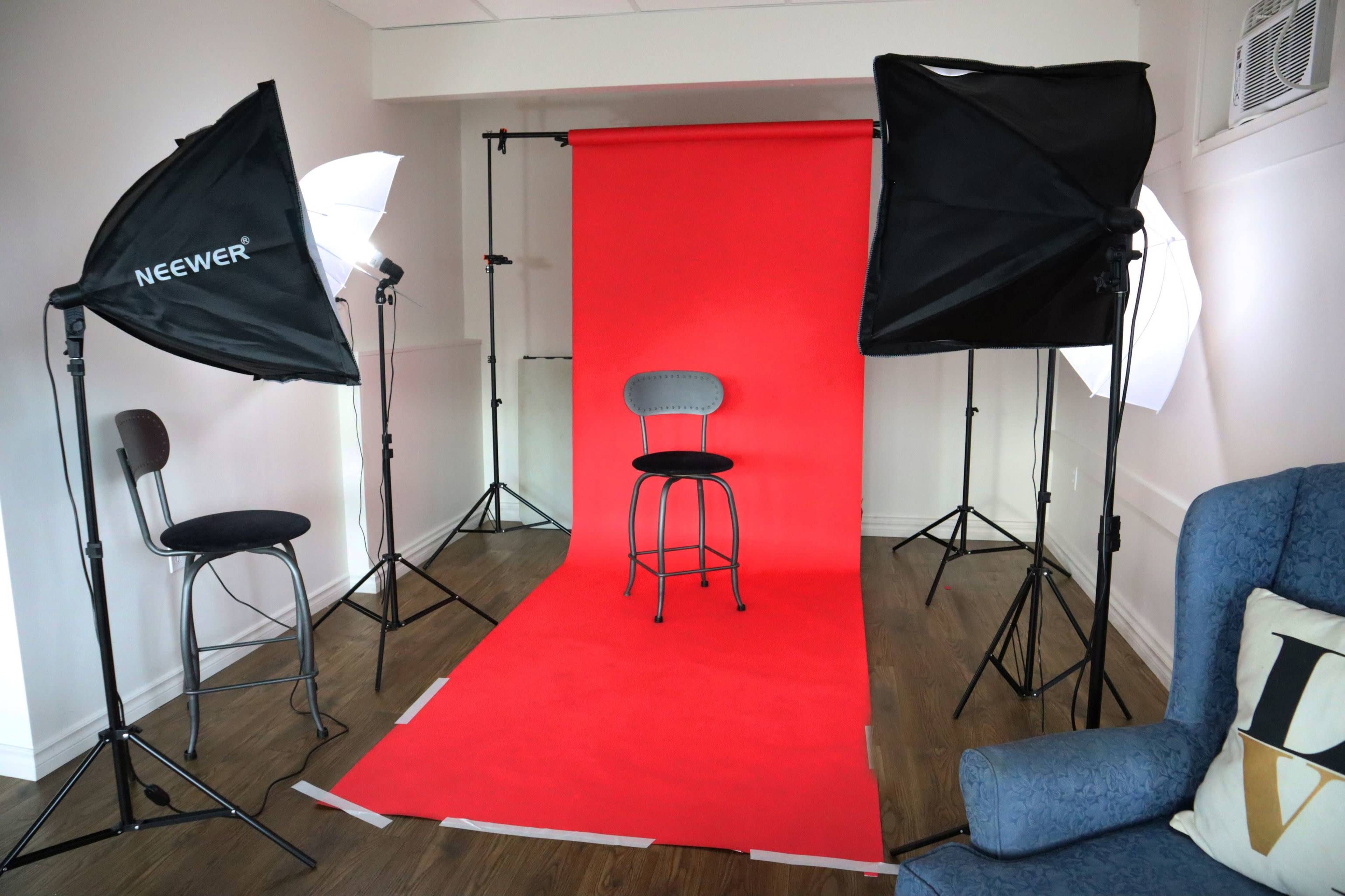 A photo studio set up with a red backdrop, three lighting setups, and a single black stool in the center.
