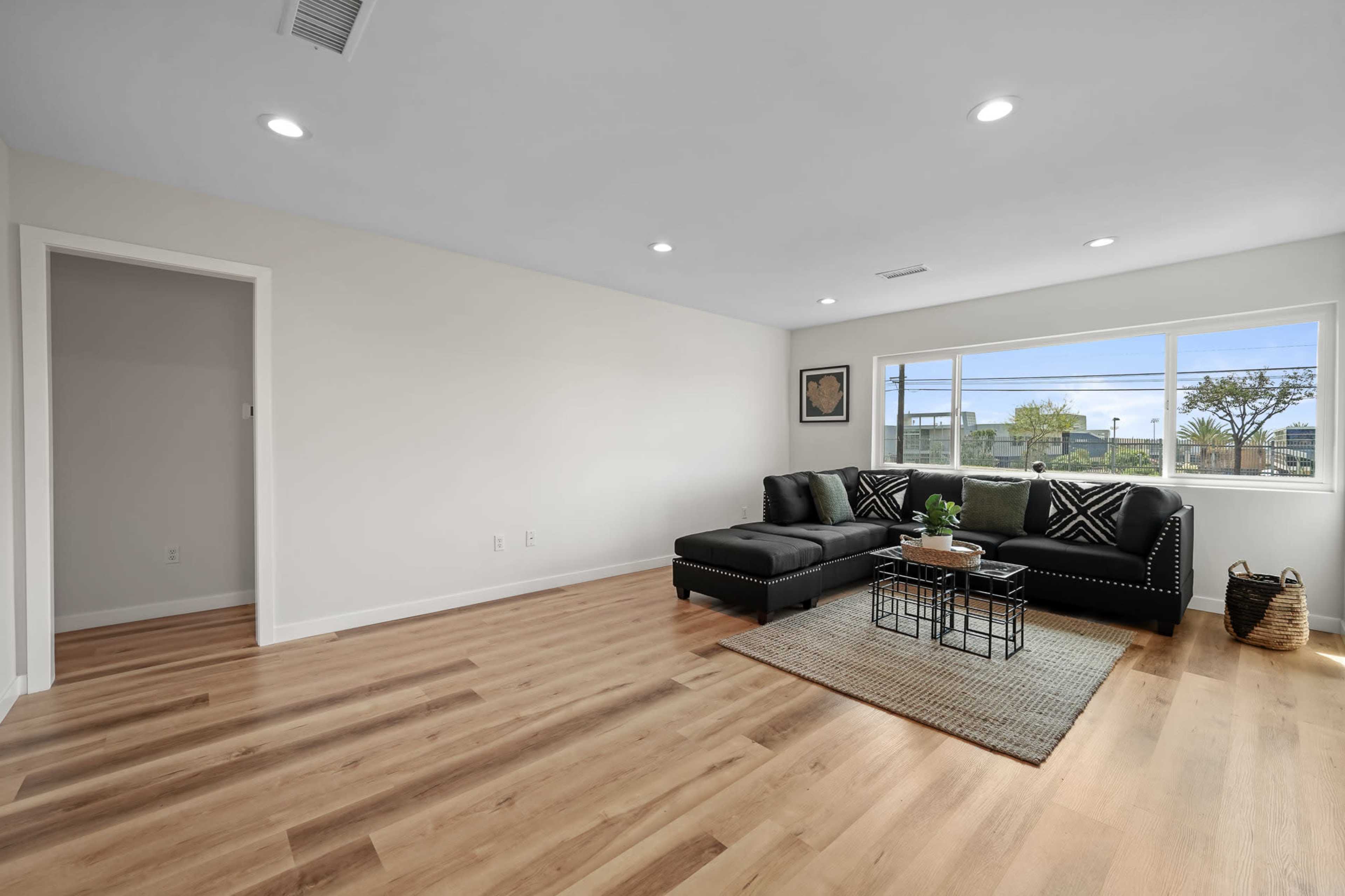 A modern living room with a black sectional sofa, a coffee table, and large windows that allow natural light to fill the space.