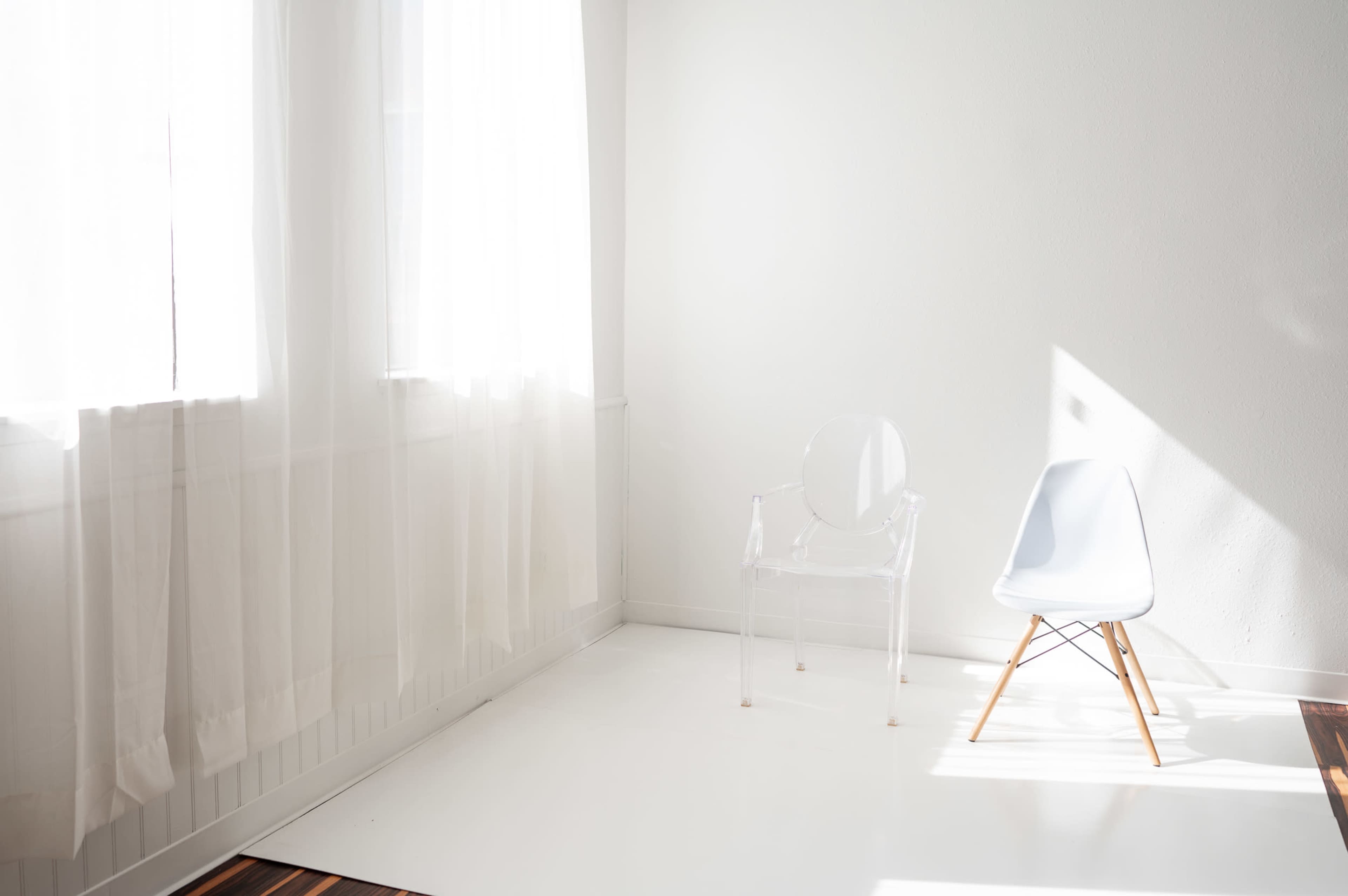 A bright, minimalist room features a clear acrylic chair and a light blue Eames chair on a white floor with sheer curtains filtering sunlight.