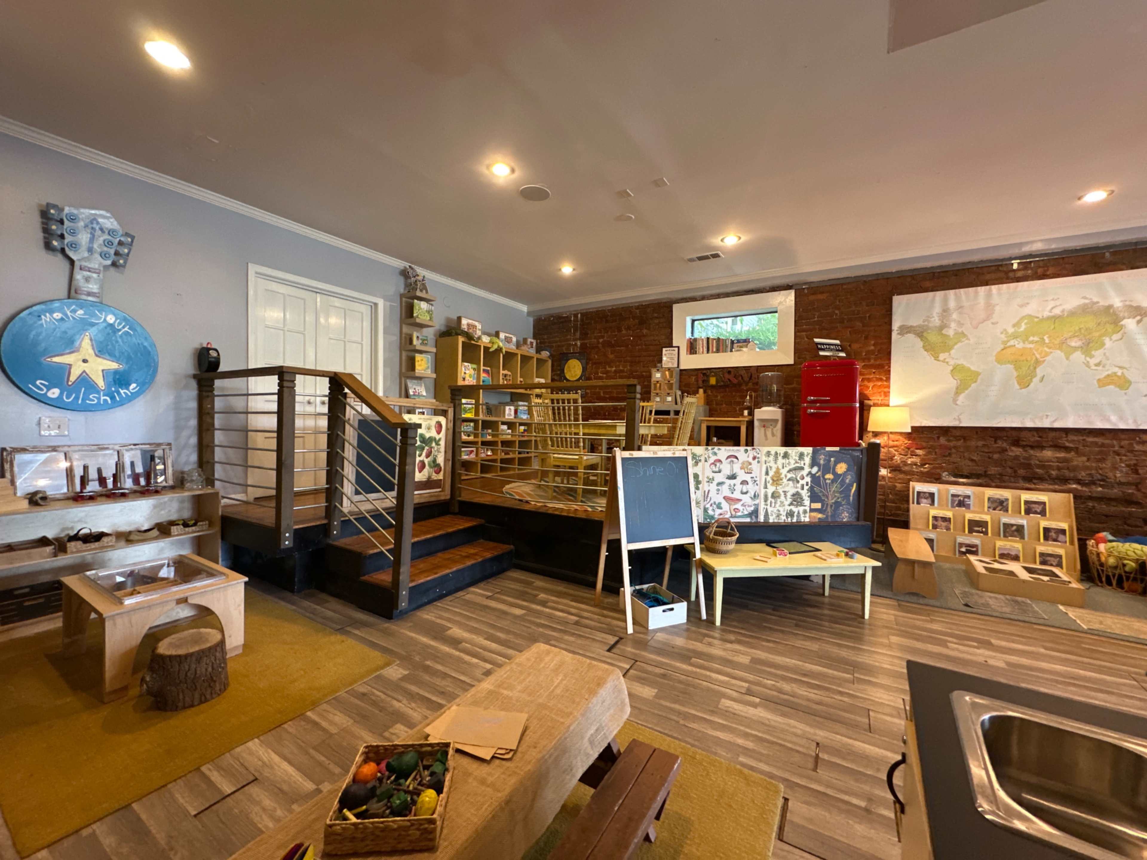 The image shows a brightly lit room with wooden flooring, featuring a play area with shelves stocked with toys, a staircase, a map on the wall, and various learning materials arranged on tables.