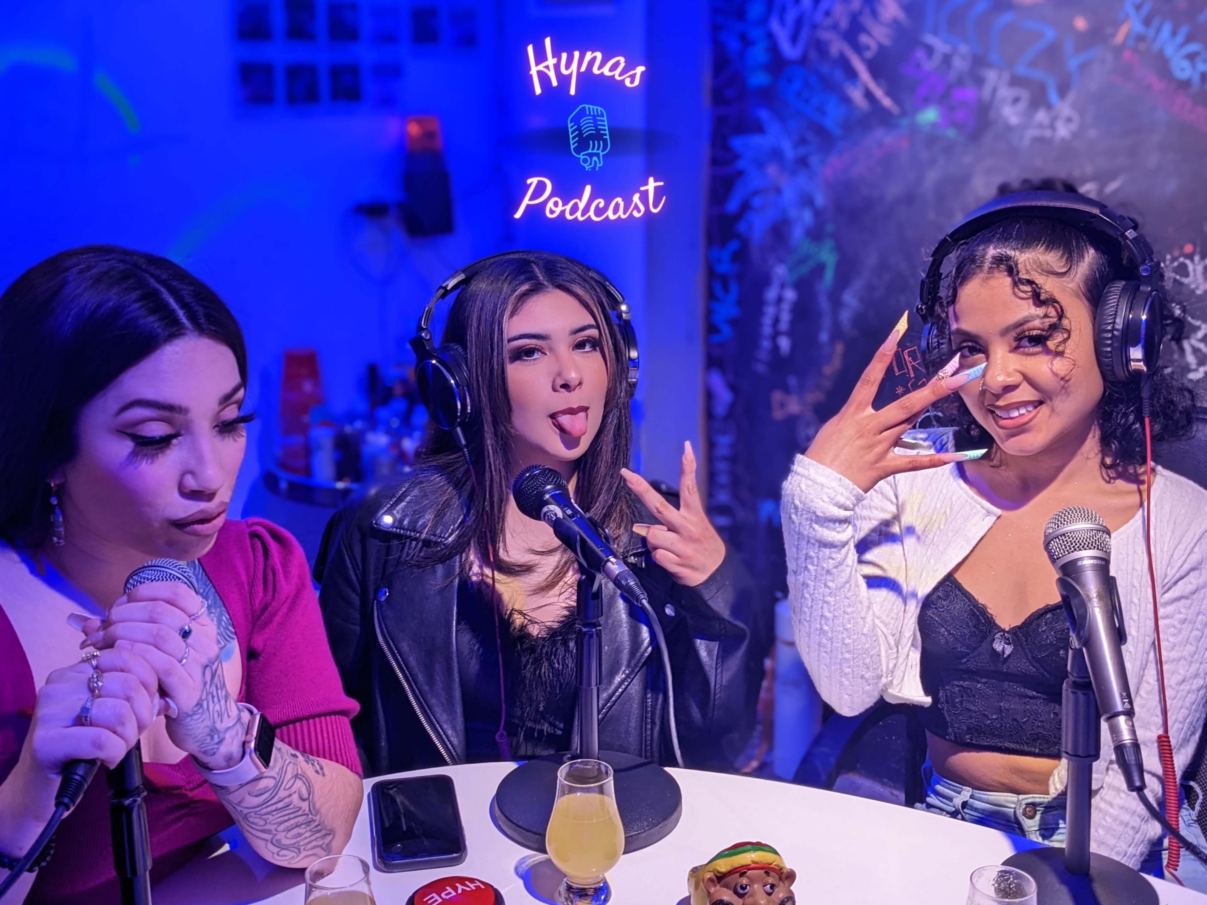 Three women sit in front of microphones in a podcast studio, with one making a playful gesture and another sticking out her tongue, while colorful lighting sets the background.