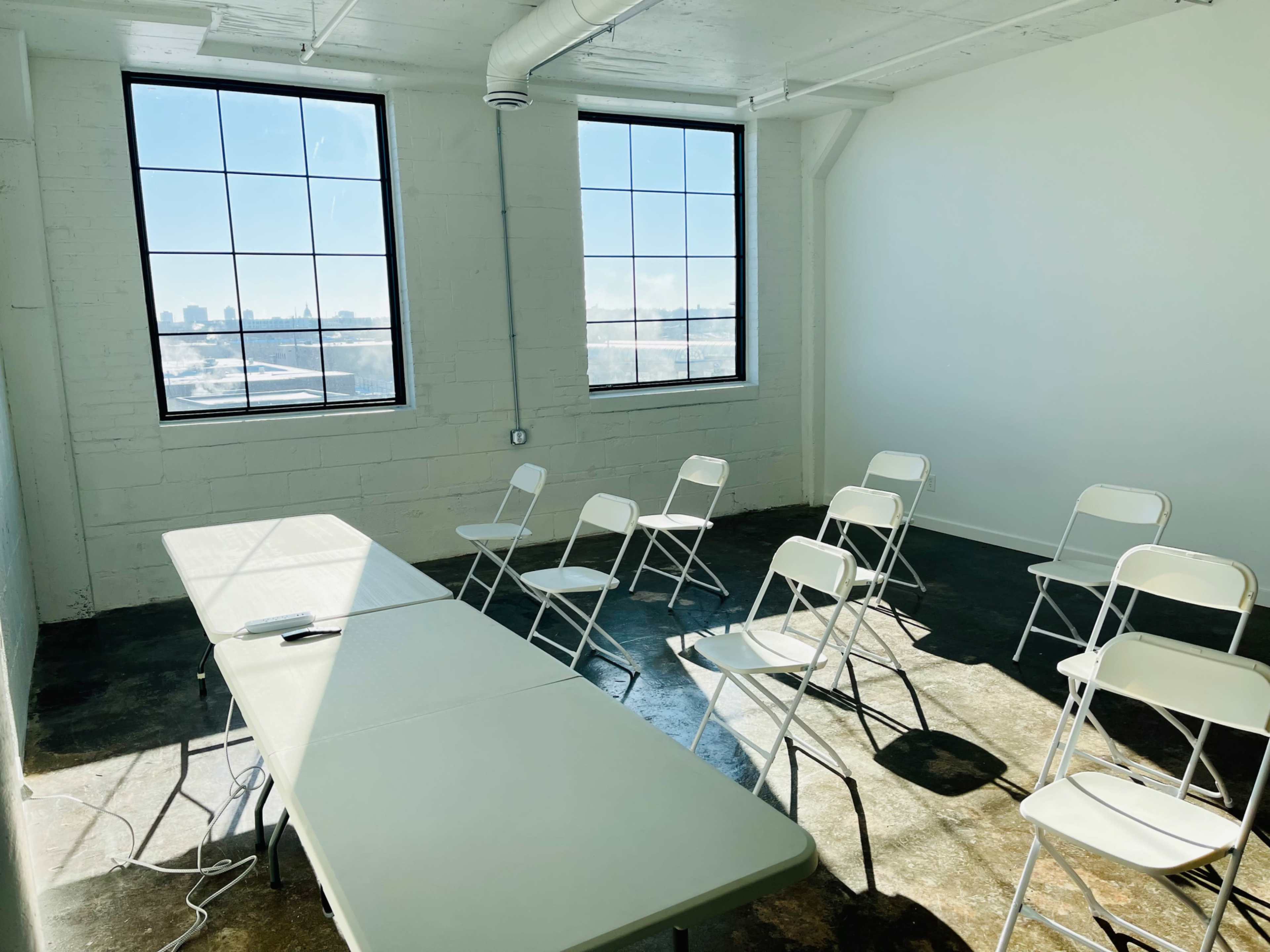 A conference room with a long table and several folding chairs arranged around it, featuring large windows that allow natural light to enter.