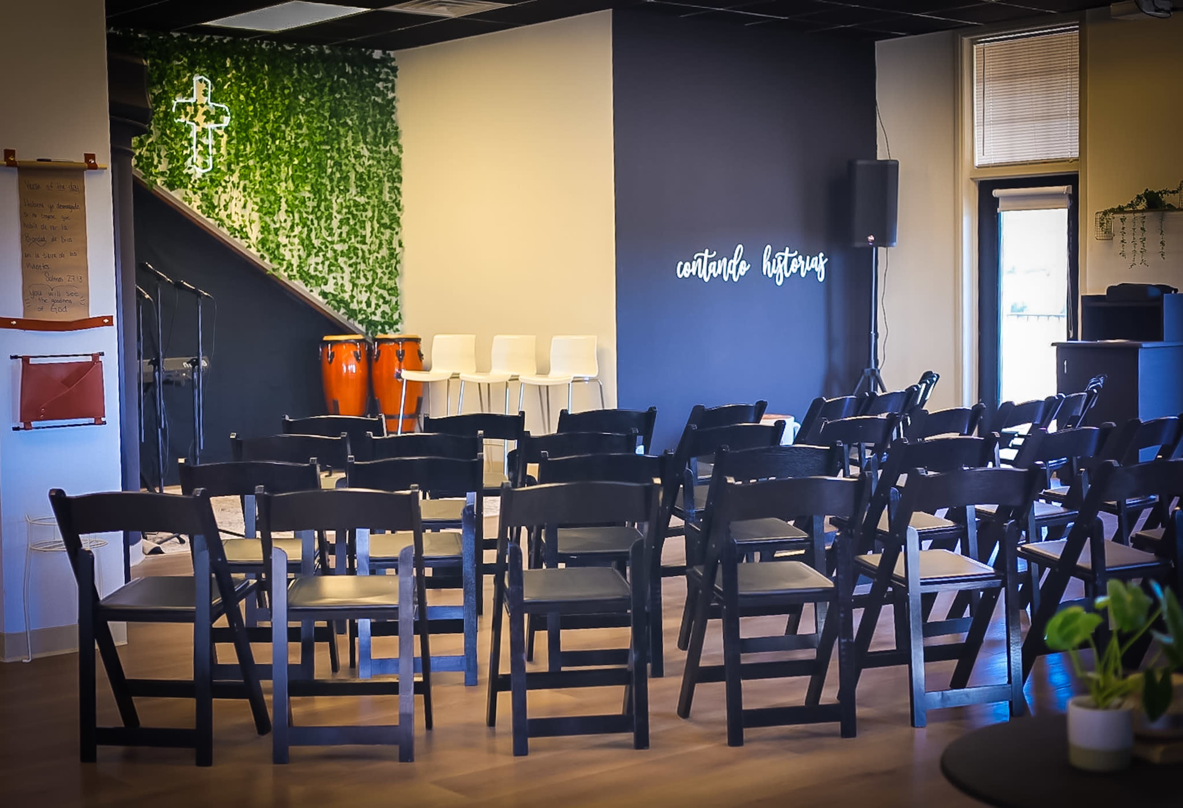 The image shows a modern event space set up with rows of black chairs facing a small stage, accompanied by instruments and greenery on the walls.