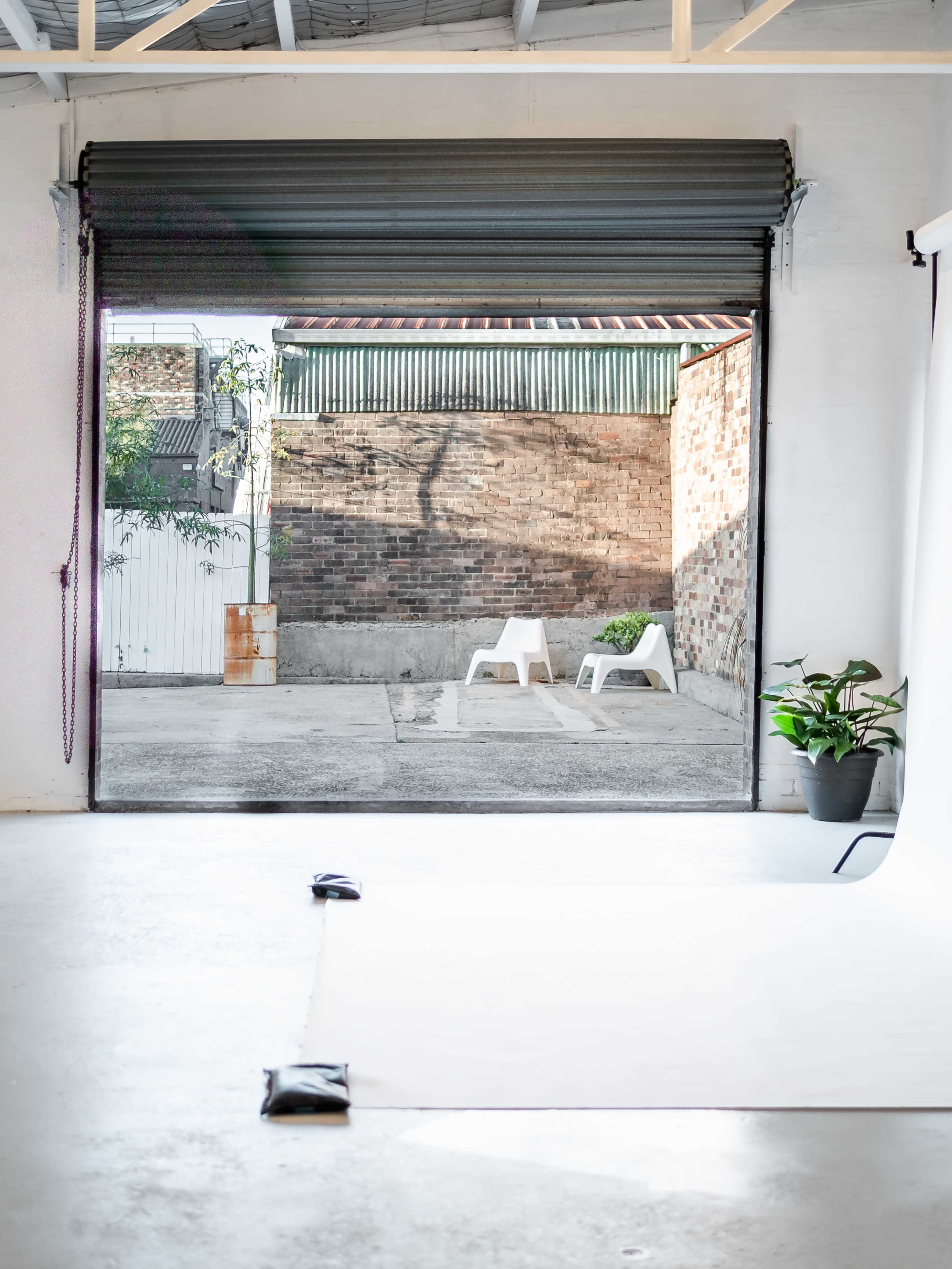 A view of a minimalist studio with an open garage door leading to a courtyard featuring a brick wall and two white chairs.