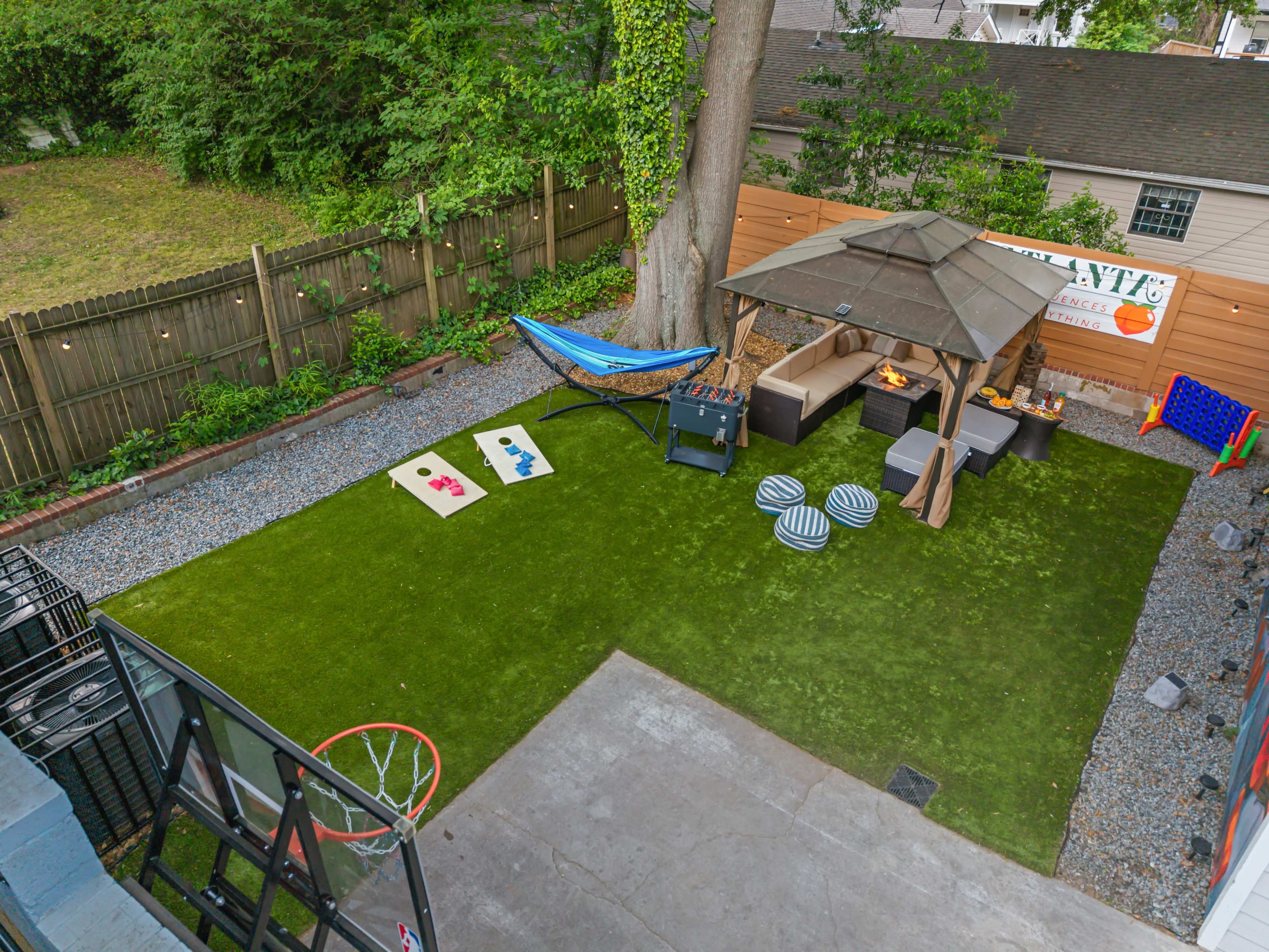 The image shows a well-maintained backyard featuring a green lawn, a shaded seating area with a fire pit, a hammock, and a small basketball hoop.