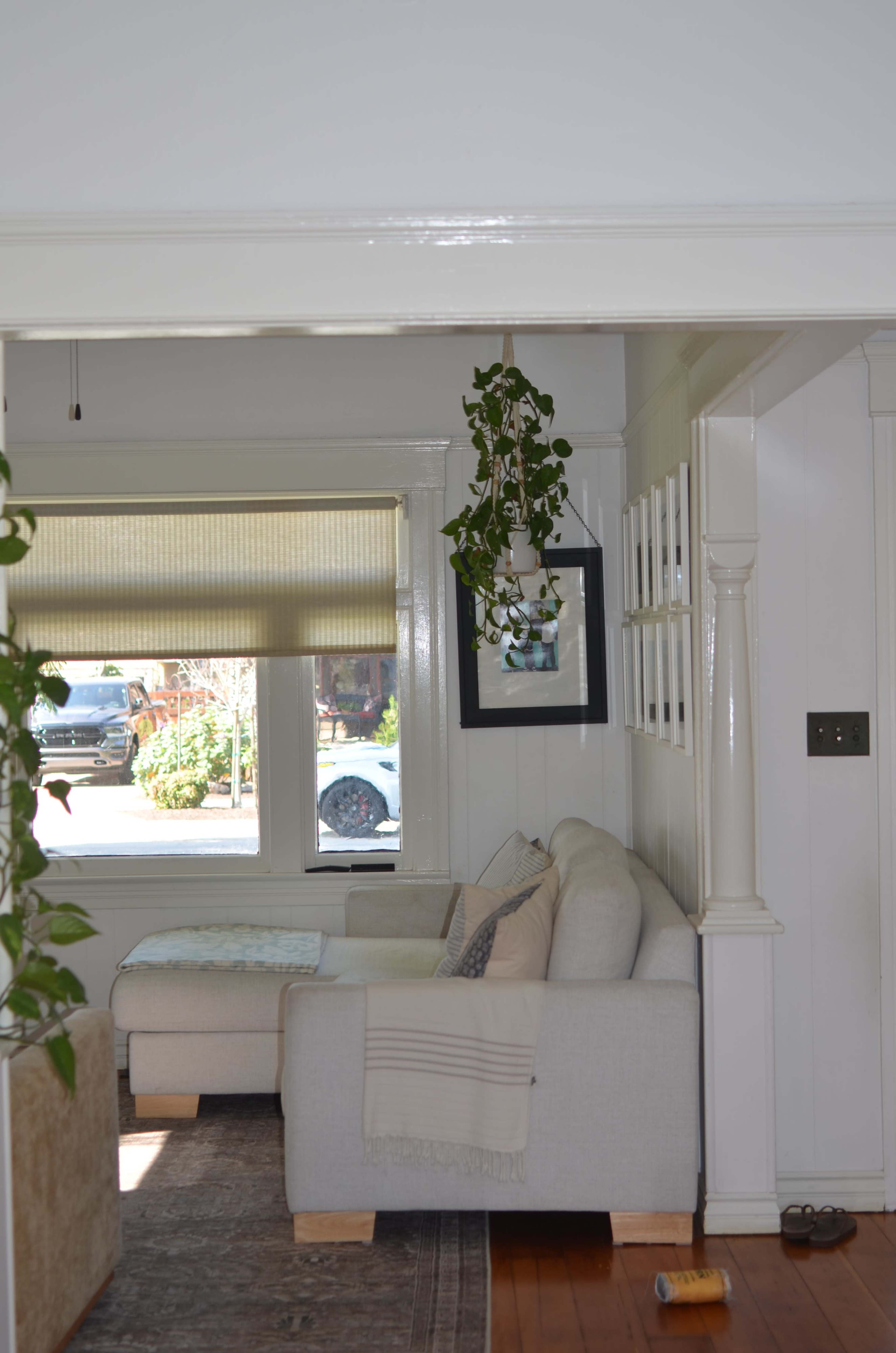 The image shows a bright living room with a white couch, decorative pillows, and a hanging plant, along with a view of a window through an open doorway.