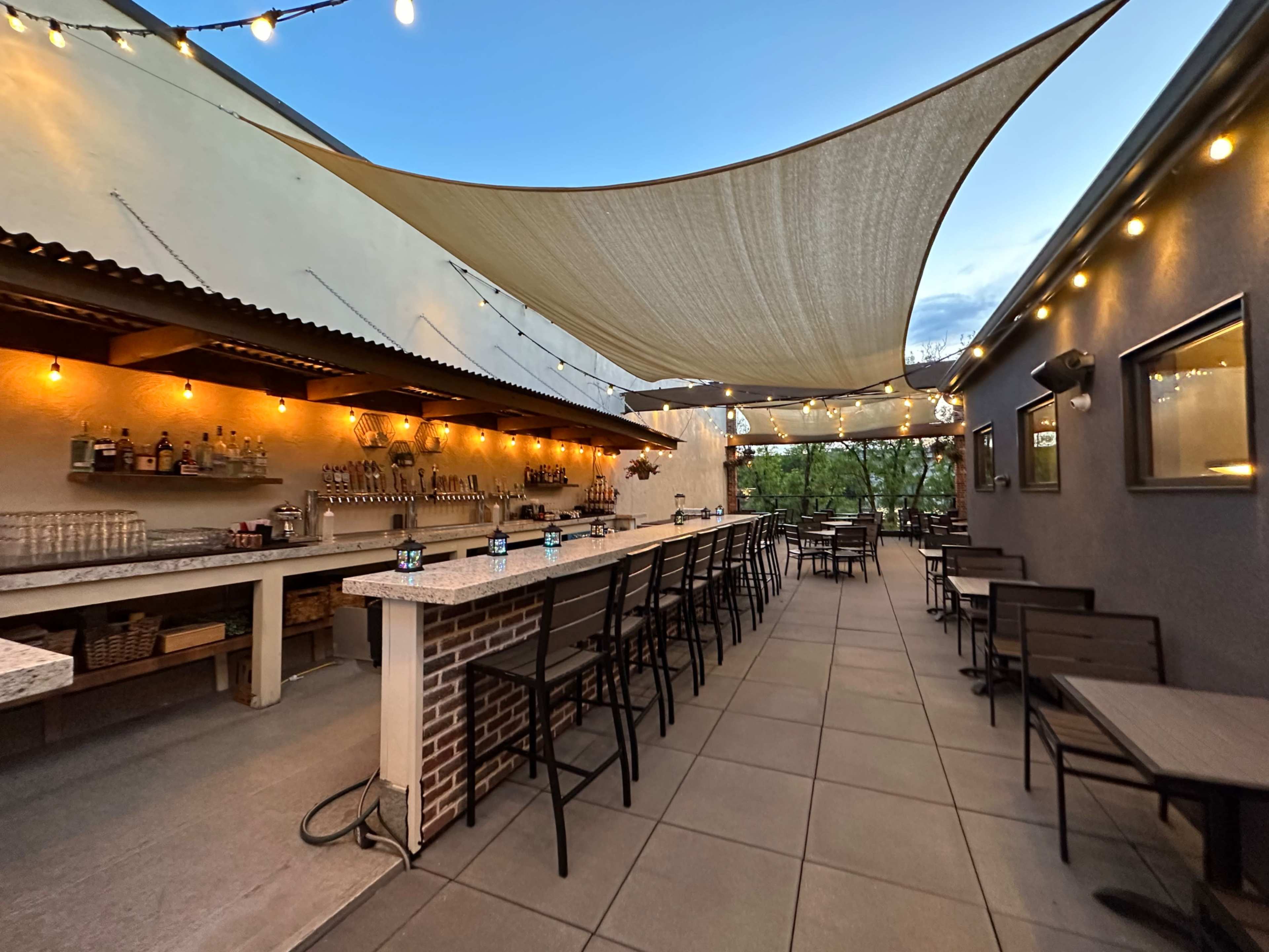 The image shows an open-air bar area with a long counter, tables, and string lights overhead, under a large canopy for shade.