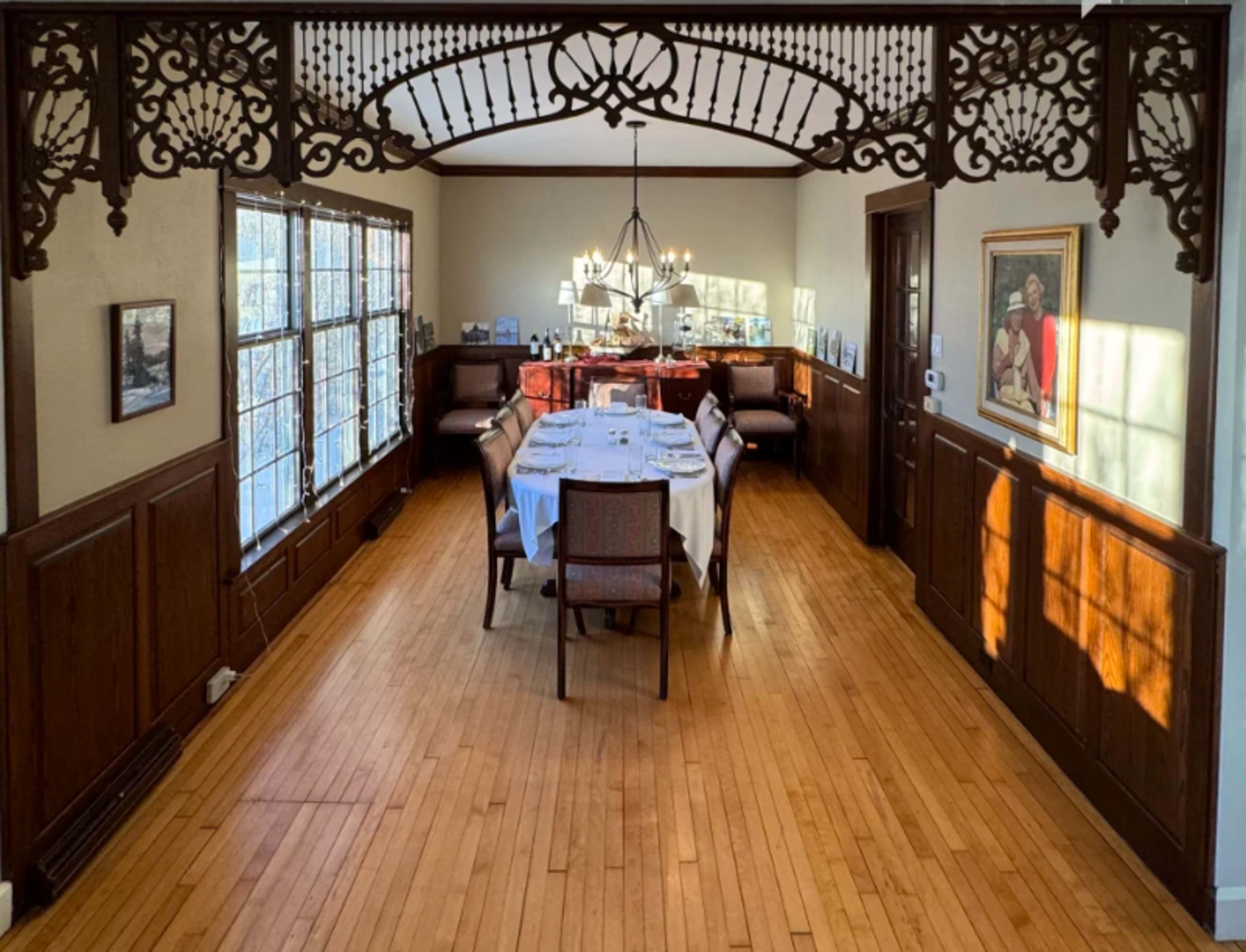 A dining room features a wooden table set for a meal, surrounded by chairs, with large windows allowing natural light to illuminate the space.