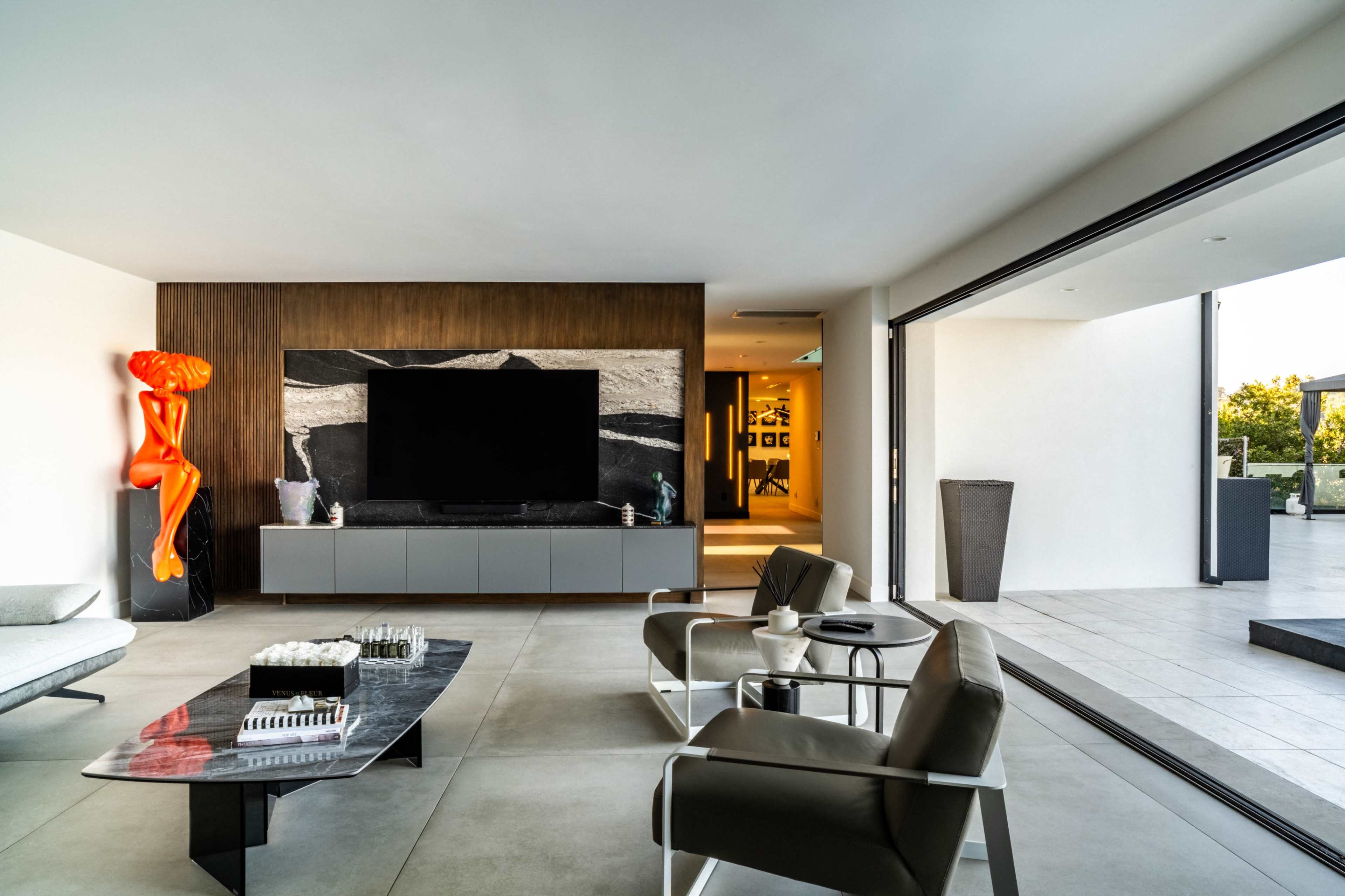 A modern living room features a large television mounted on a wooden wall with an abstract sculpture nearby, and large sliding glass doors that open to an outdoor space.
