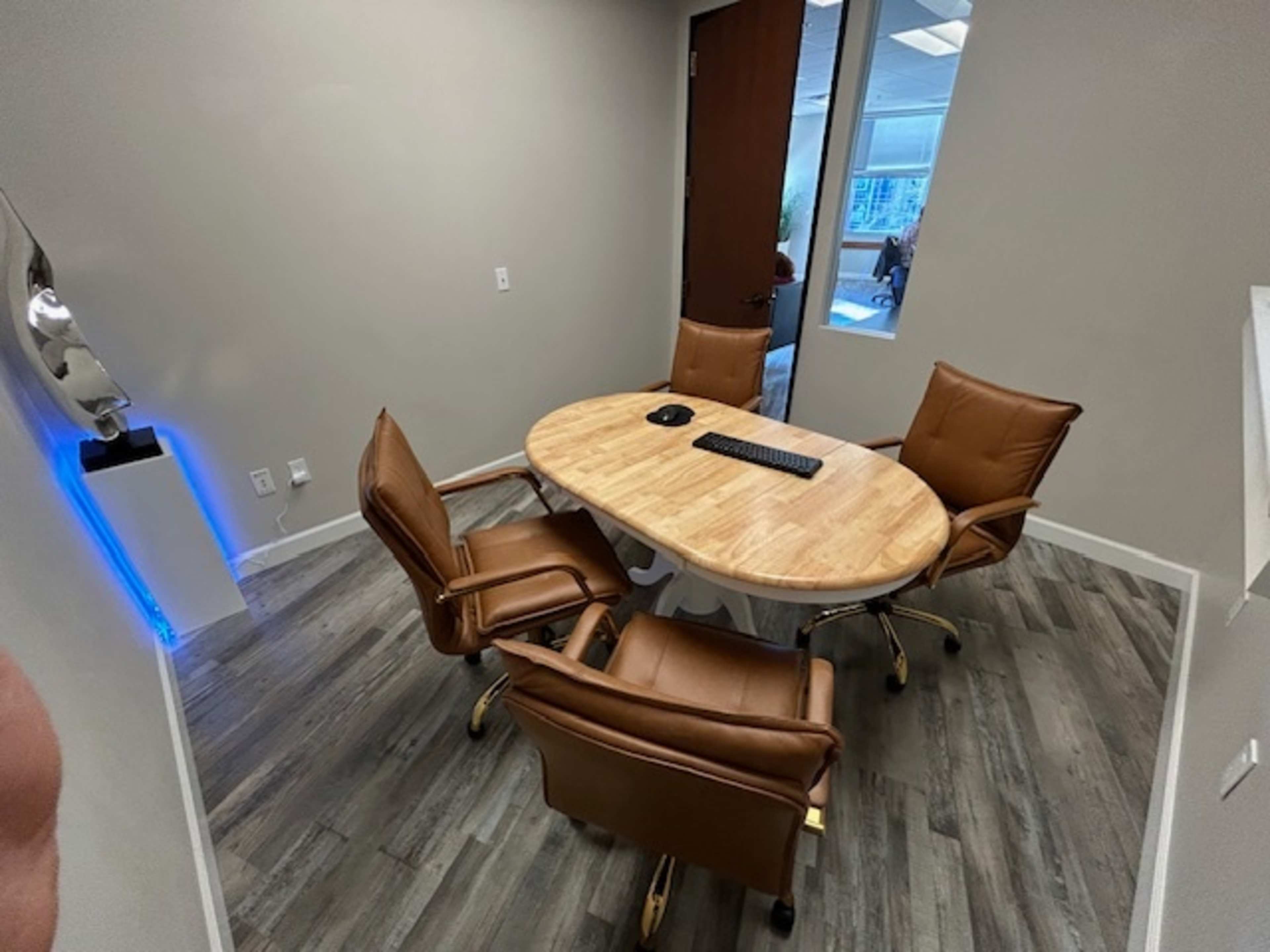 A conference room features a round wooden table surrounded by four brown leather chairs, with a keyboard on the table and a glass door in the background.