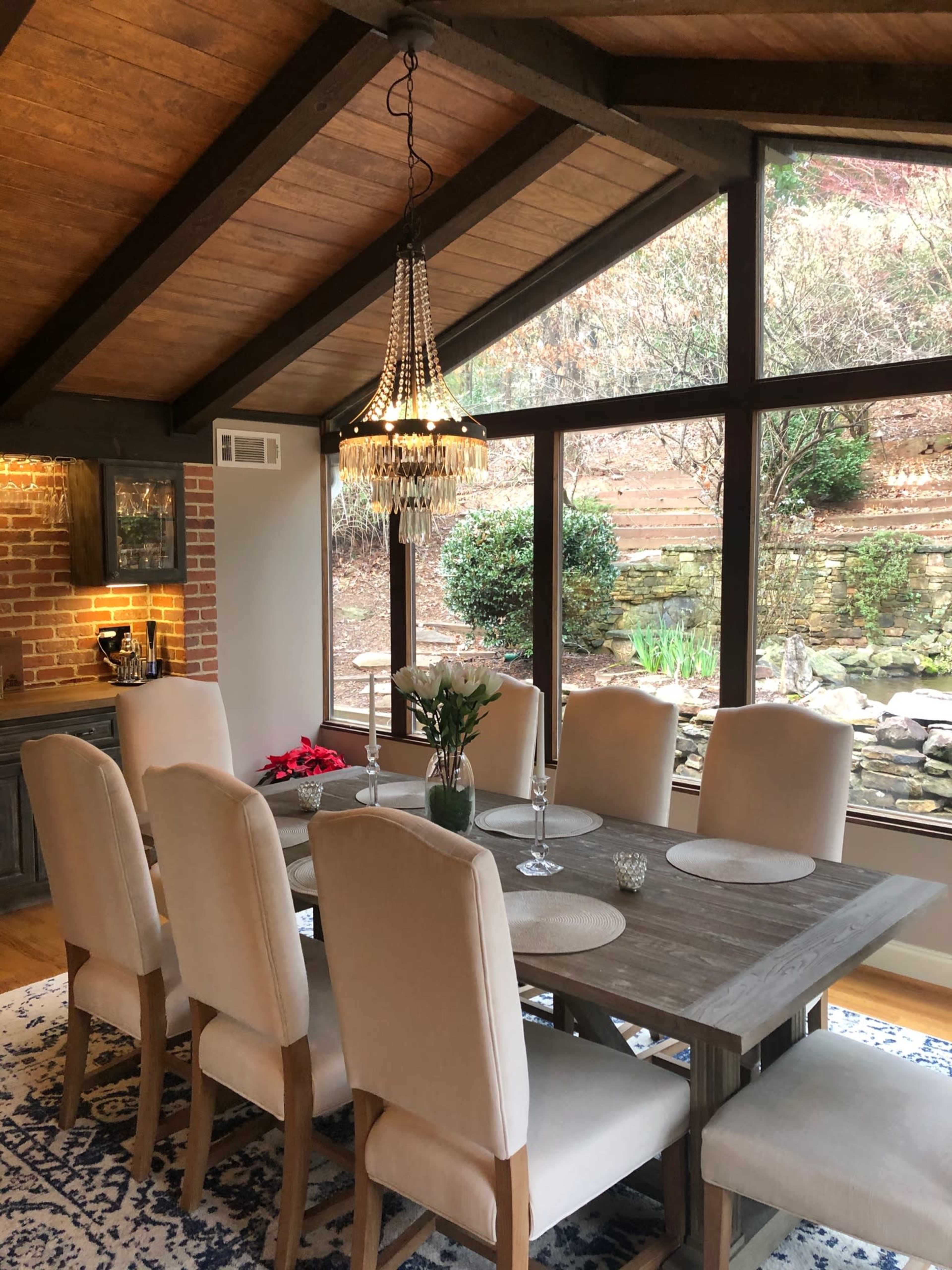 The image shows a spacious dining area with a wooden table surrounded by upholstered chairs, a chandelier above, and large windows offering a view of a landscaped outdoor area.