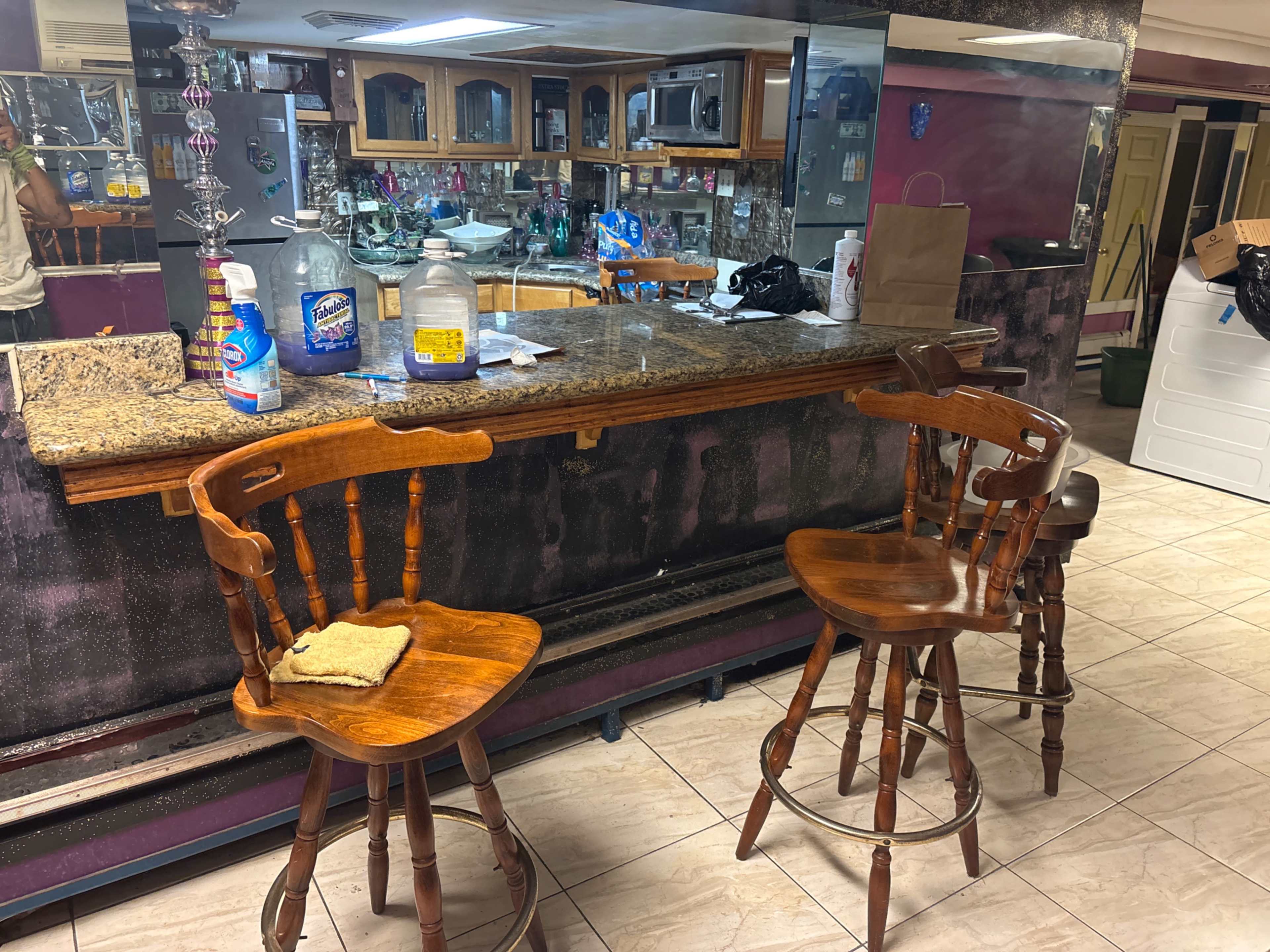 The image shows a bar area with two wooden stools, a granite counter topped with various cleaning supplies and containers, and a mirrored wall in the background.