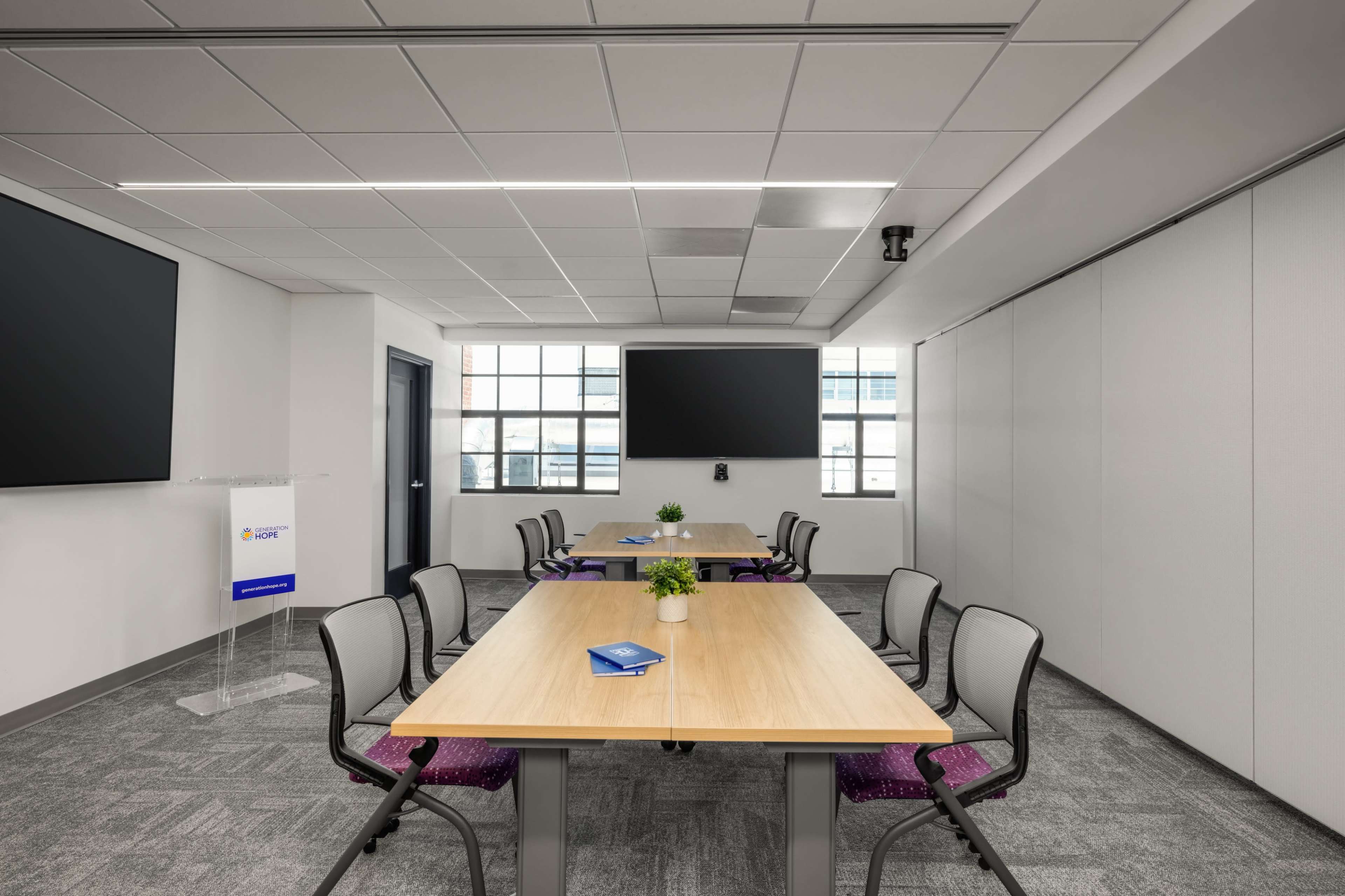 The image shows a modern conference room with two large screens, several tables arranged for meetings, and ergonomic chairs, all surrounded by large windows.