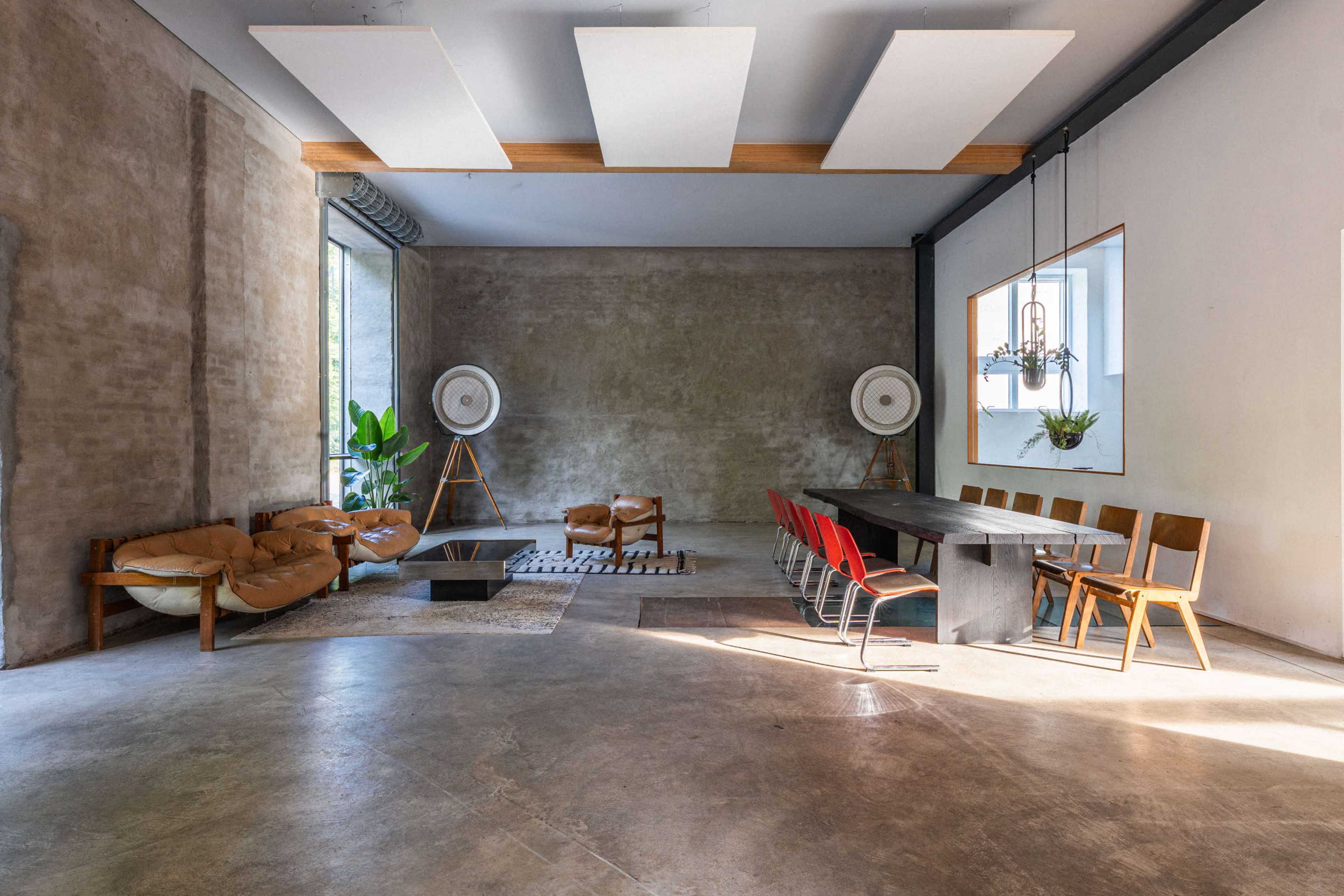 The image shows a spacious, modern interior featuring a large wooden table surrounded by chairs, a leather sofa, and large windows with indoor plants.