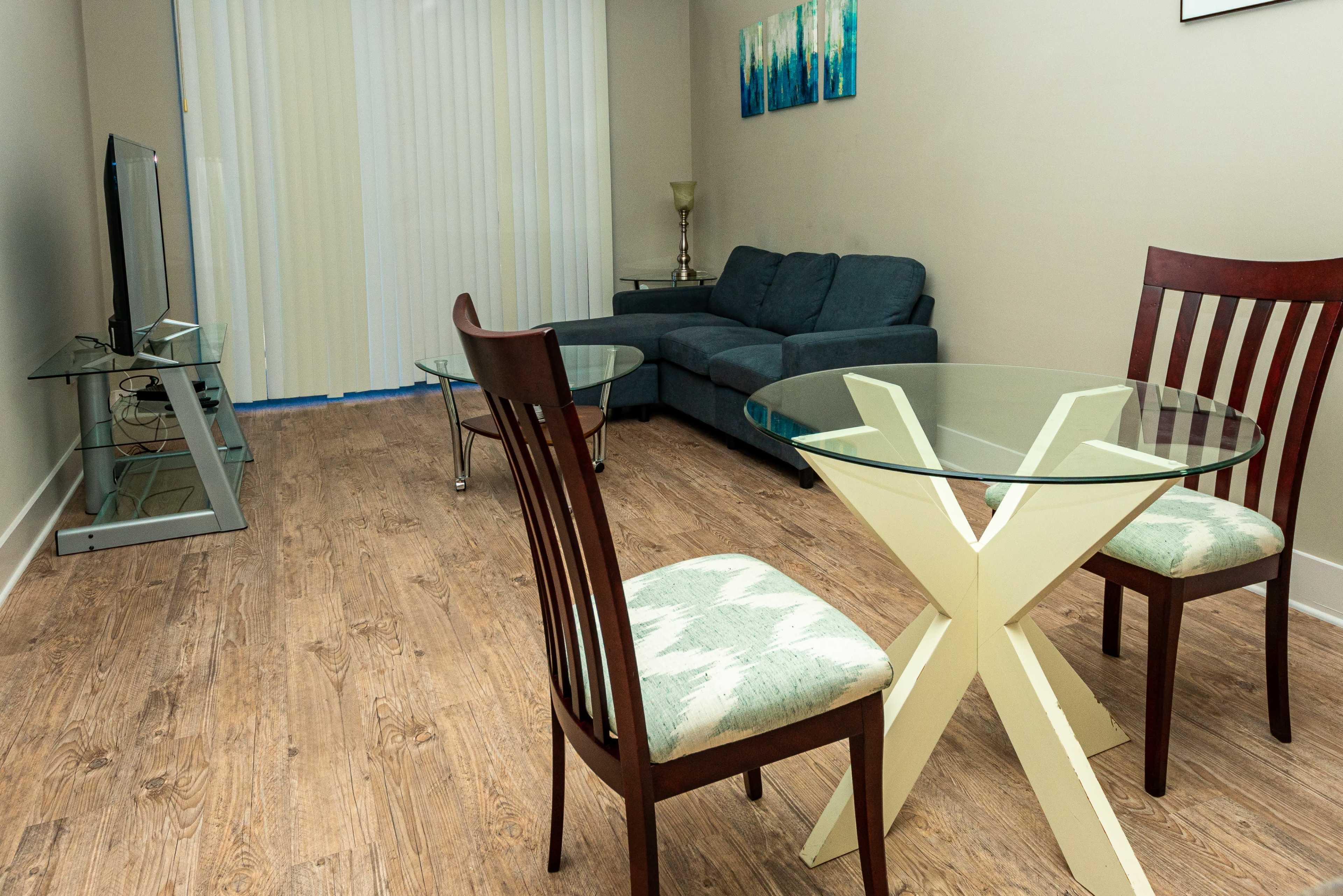 A living room area with a blue couch, a glass dining table with two wooden chairs, and a television on a glass stand.