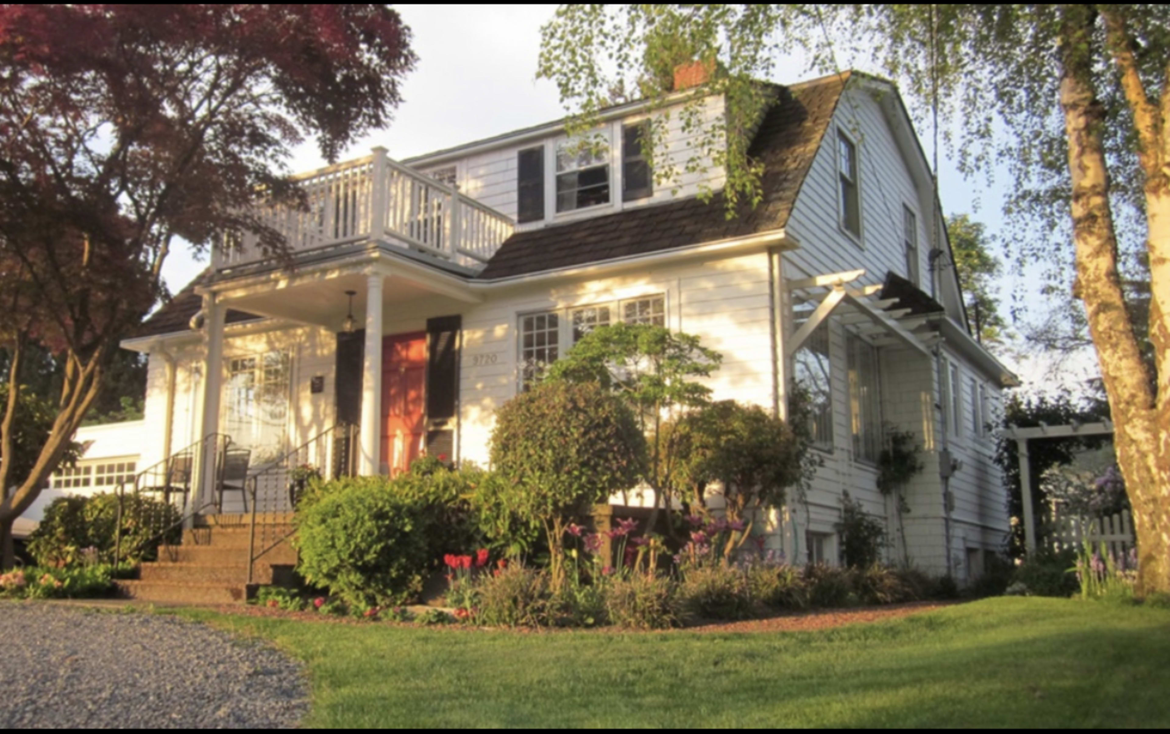 A two-story white house with a sloped roof, a front porch, and colorful garden landscaping.