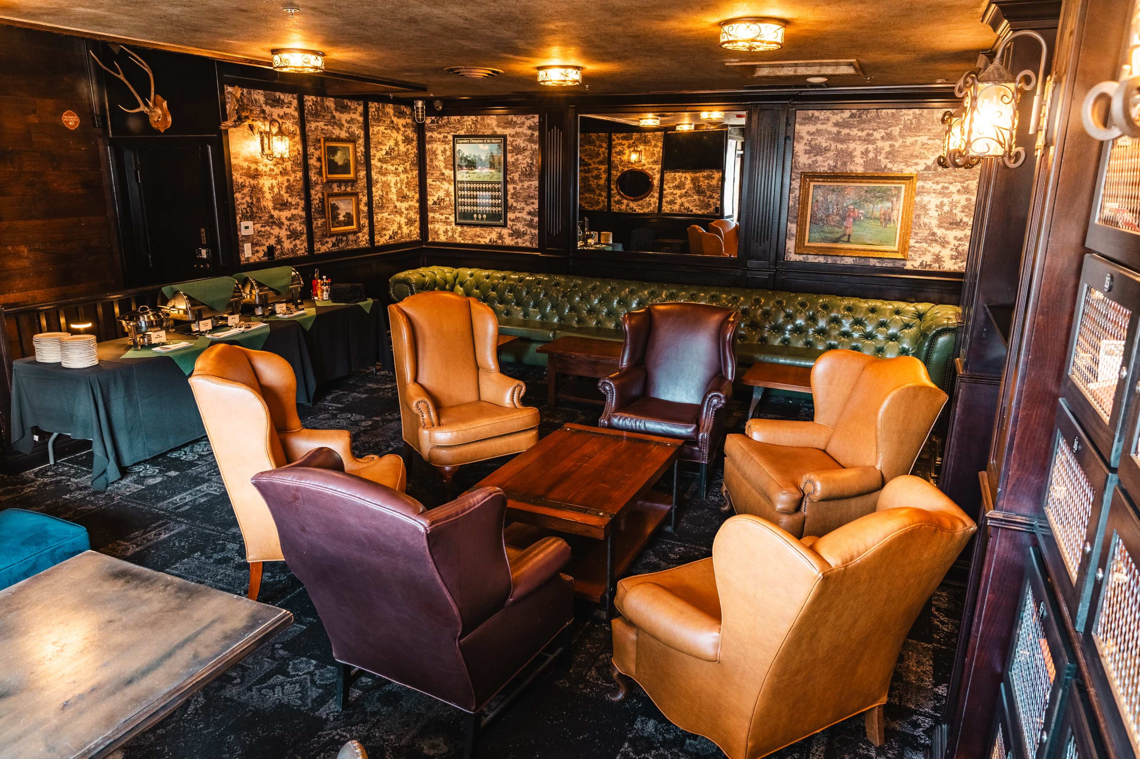 The image shows a cozy lounge area with a mix of leather armchairs, a wooden table, and patterned wallpaper.