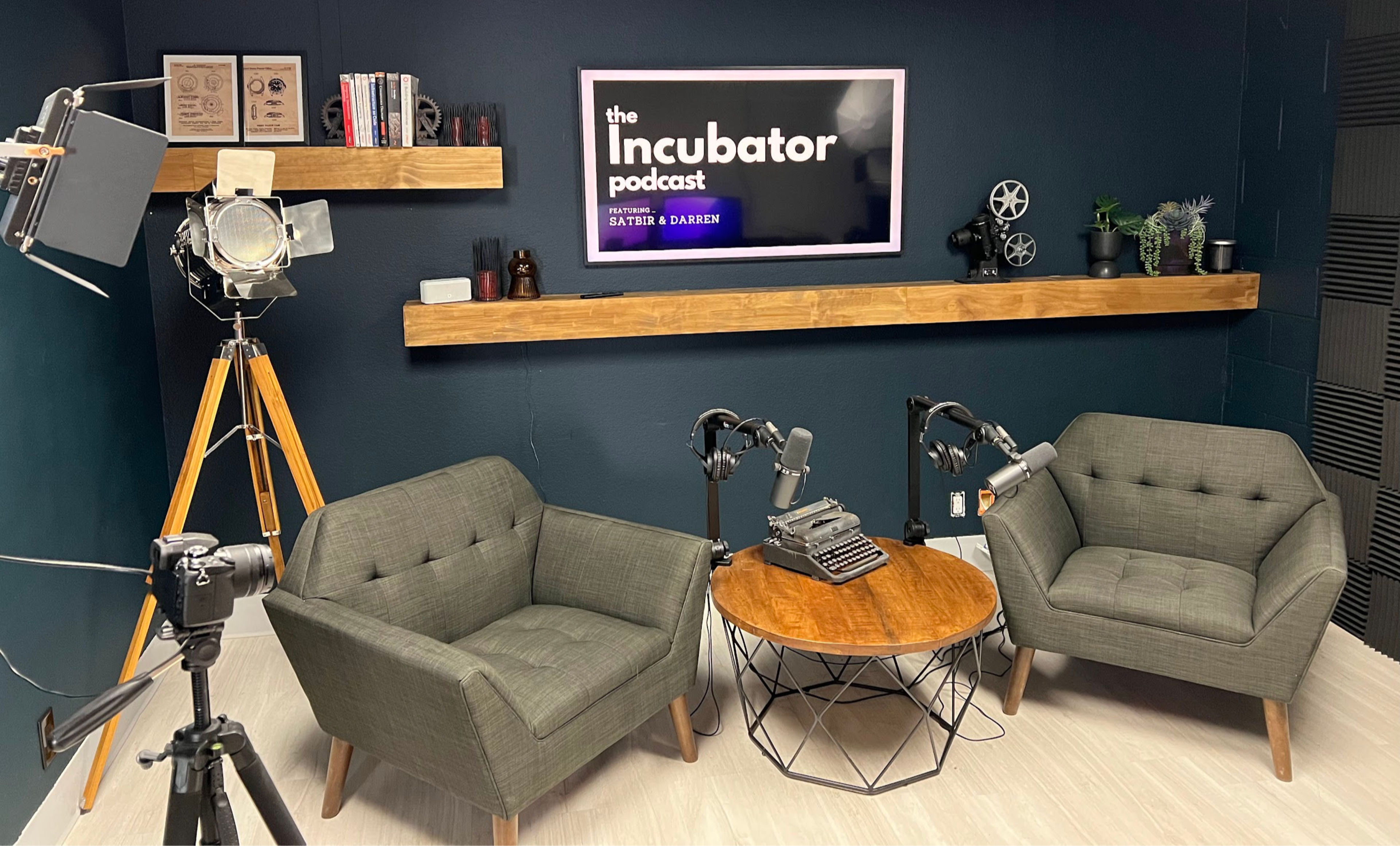 A podcast recording studio with two green armchairs, a round table, a vintage typewriter, and a television displaying the title "the Incubator podcast" on a dark blue wall.