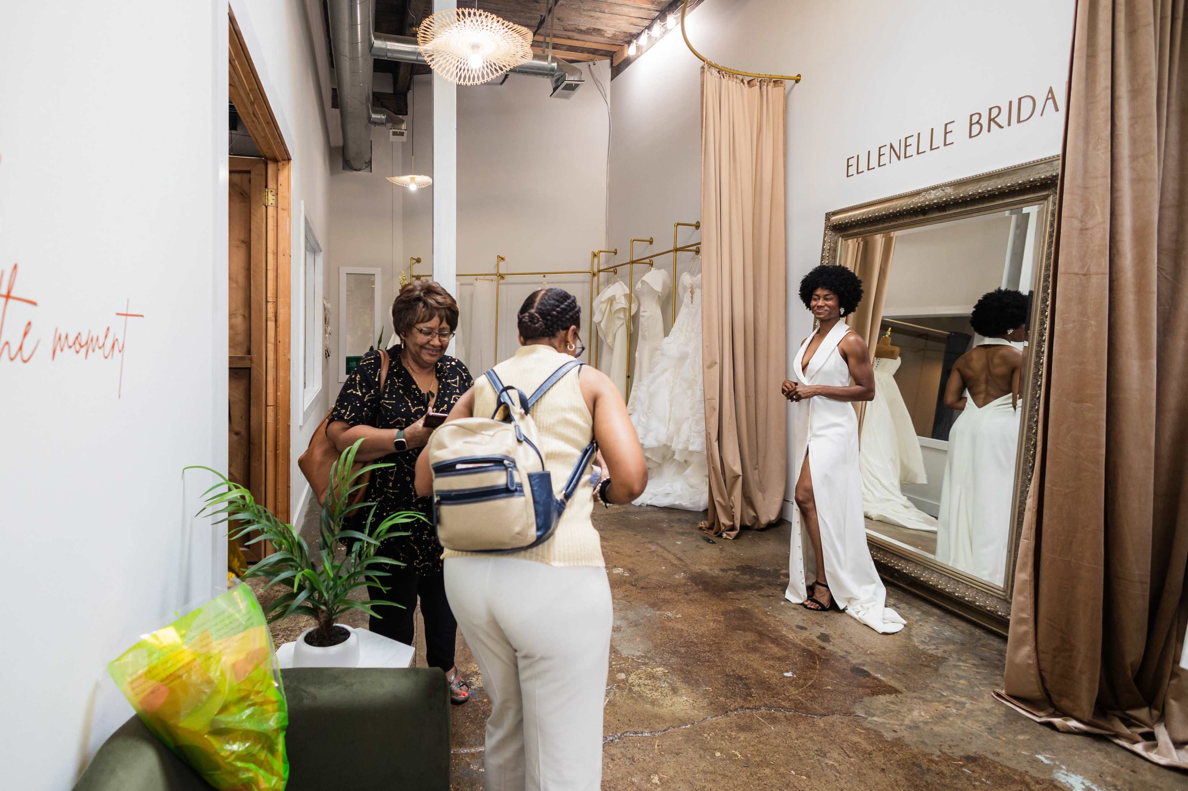 A bride stands in a fitting area while two women engage in conversation nearby, surrounded by wedding dresses and a large mirror.