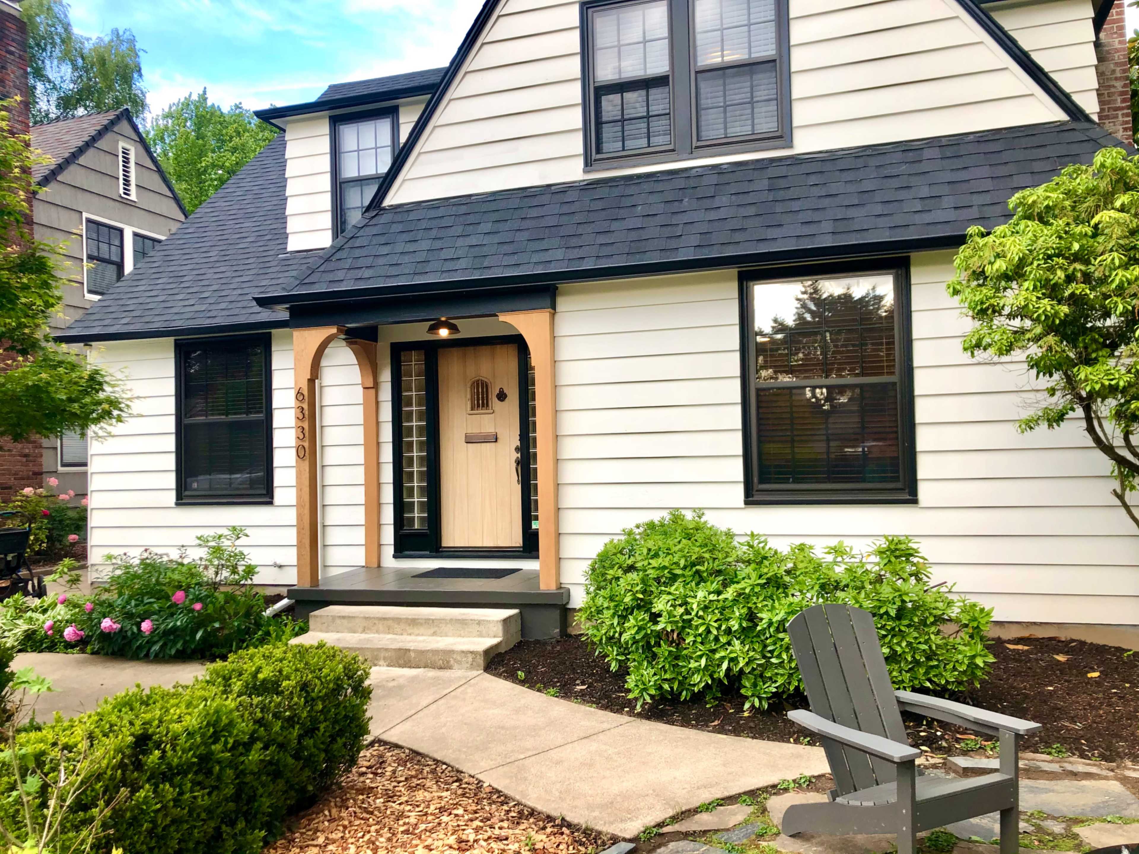 A modern, two-story house with a triangular roof, featuring a wooden front door and surrounded by well-maintained shrubs and floral beds.