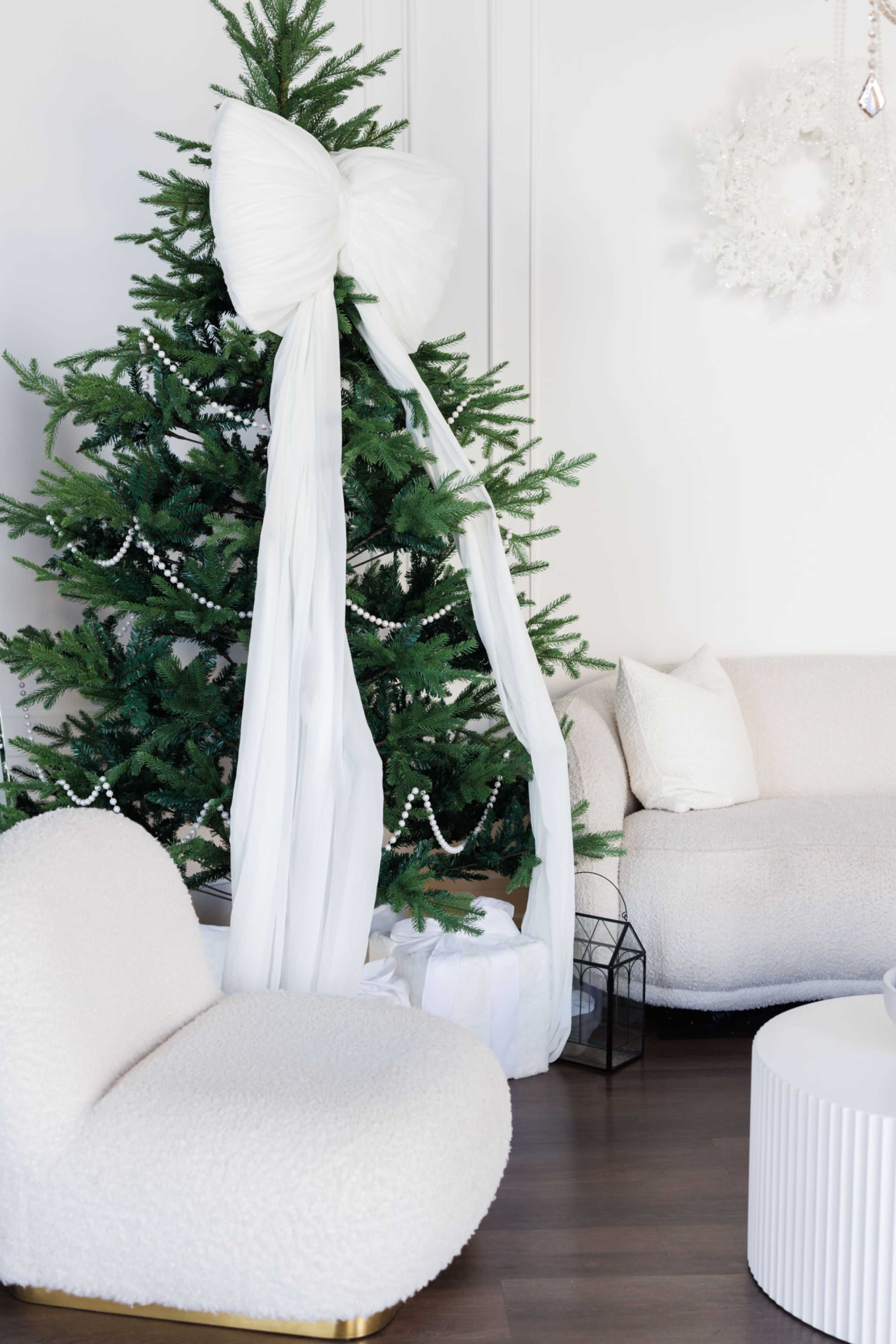 A decorated Christmas tree with a large white bow stands next to a modern, plush beige chair and a white circular table in a minimalist living room.