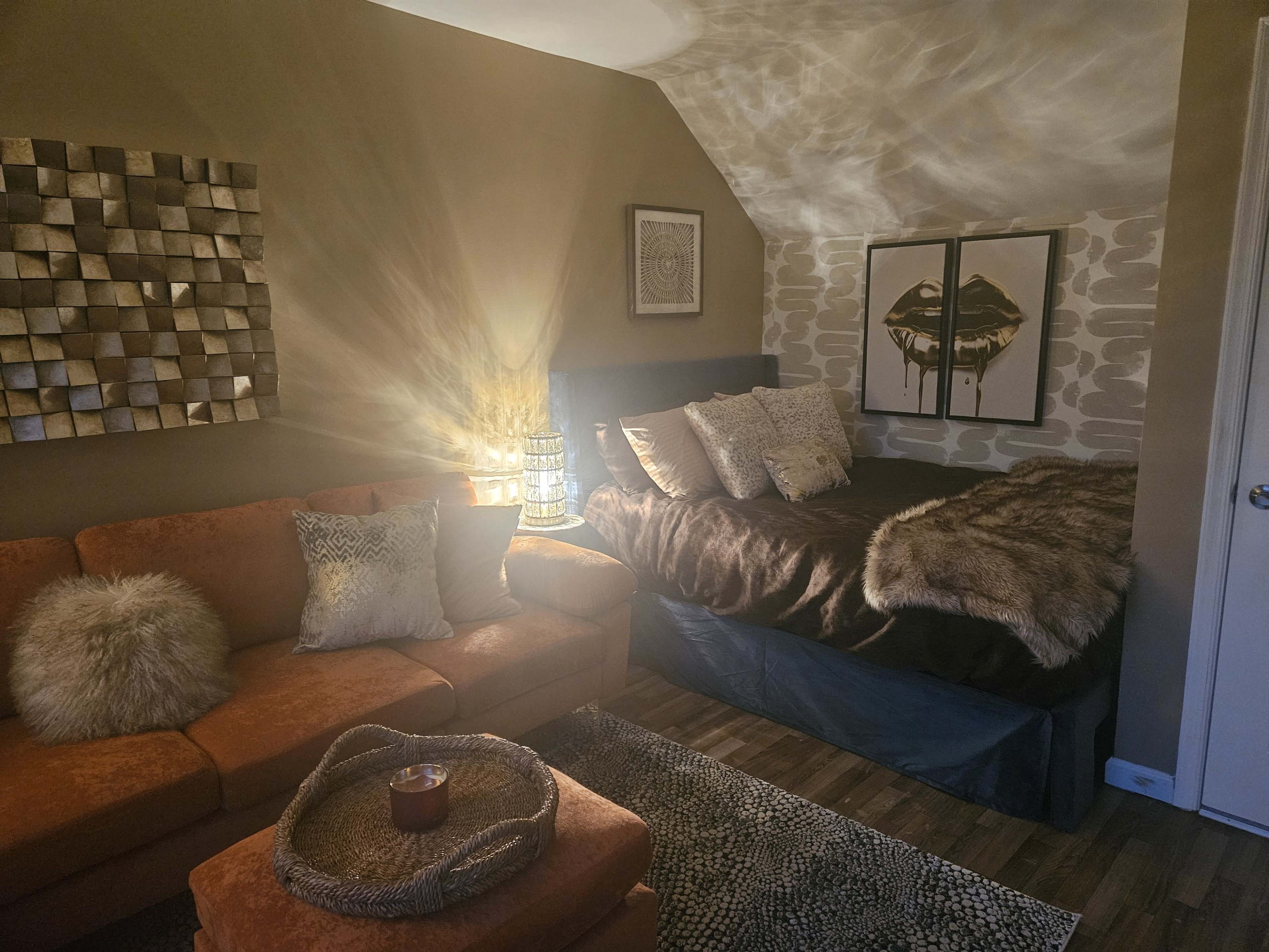 The image shows a cozy bedroom with a brown bed covered in a fur blanket, an orange couch, and decorative wall art illuminated by soft lighting.