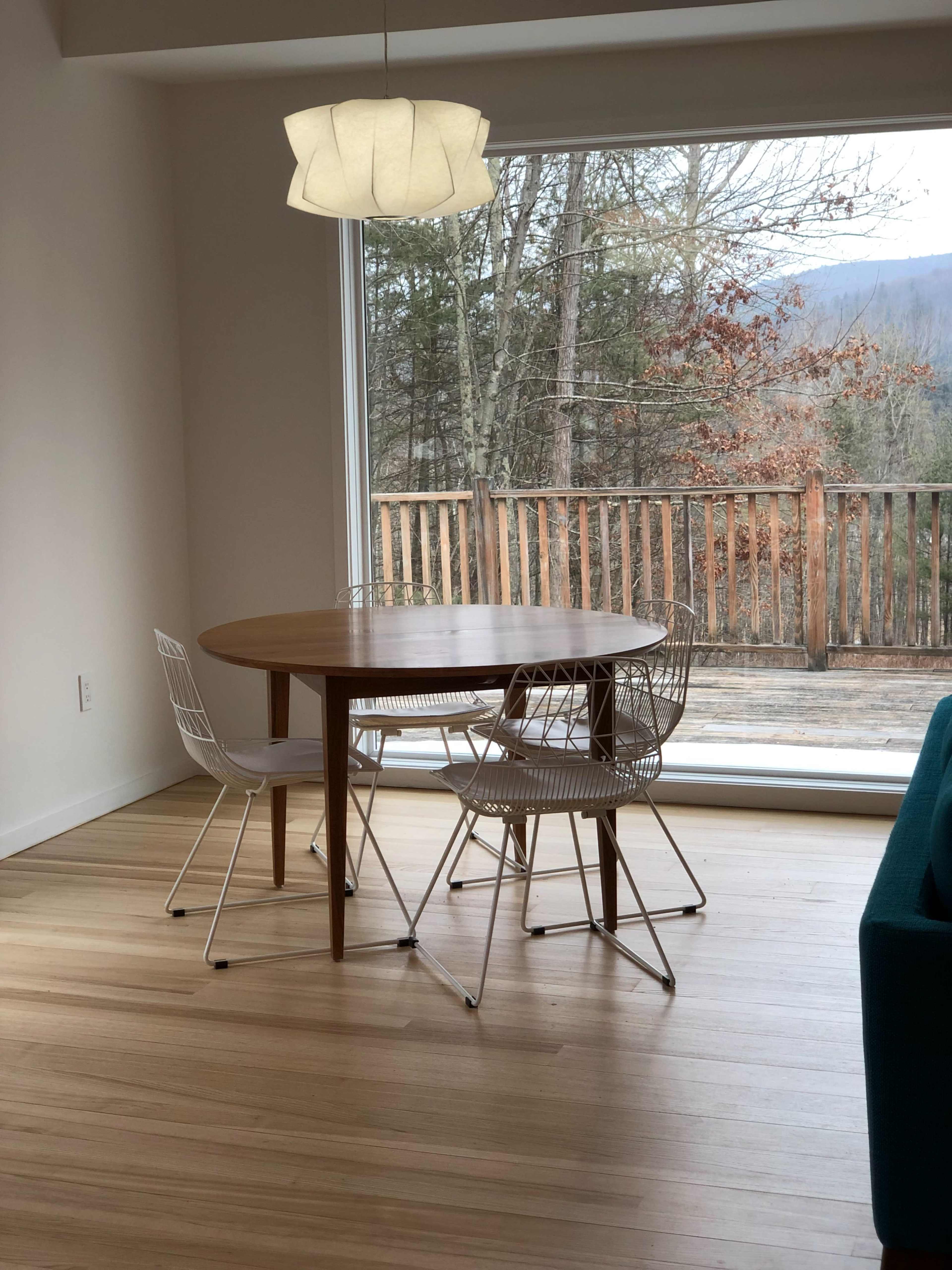 A round wooden dining table with four chairs is situated by a large window overlooking a forested area.